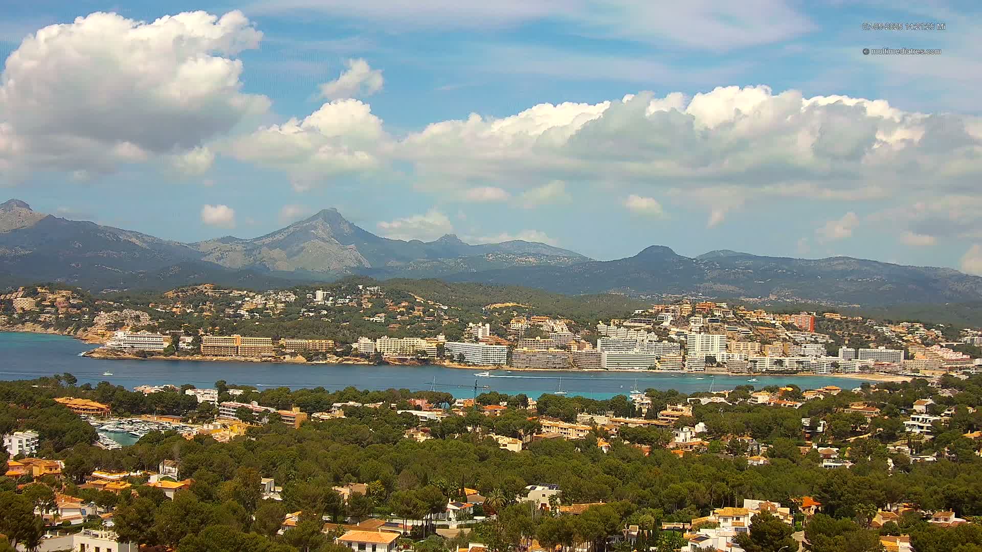A coastal town nestled beside a calm bay, backed by lush green hills and a mountain range under a partly cloudy sky.