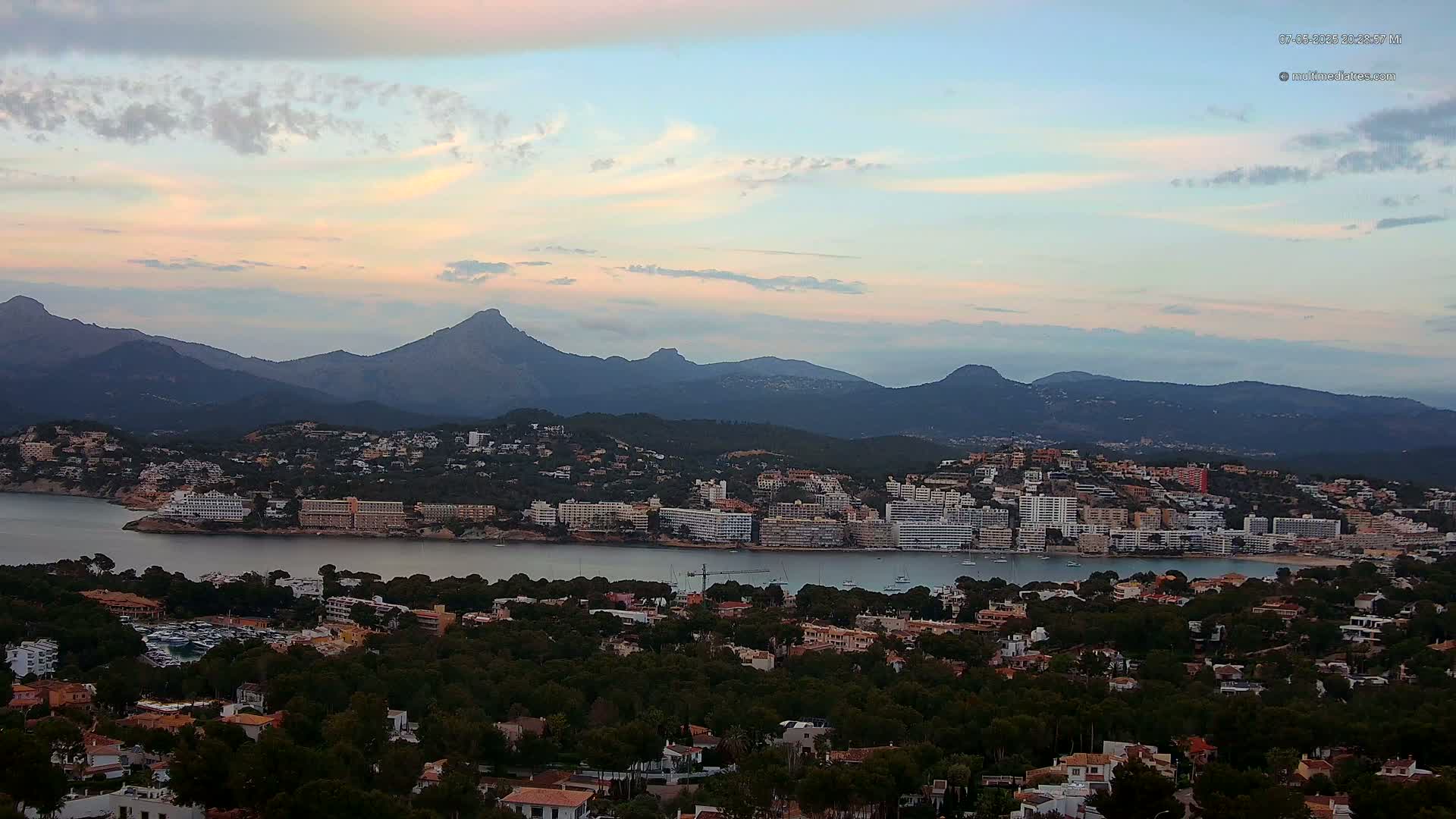 A coastal town nestled between a calm bay and a mountain range under a partly cloudy, pastel-colored sky.