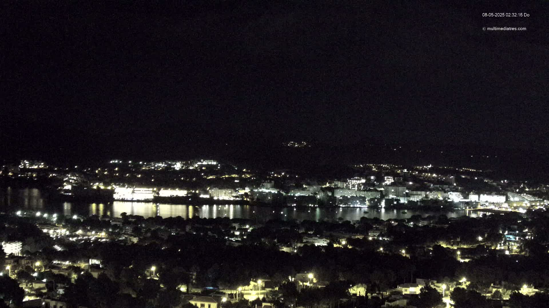 A nighttime view of a coastal town with numerous lights reflecting on a calm body of water, under a clear, dark sky.