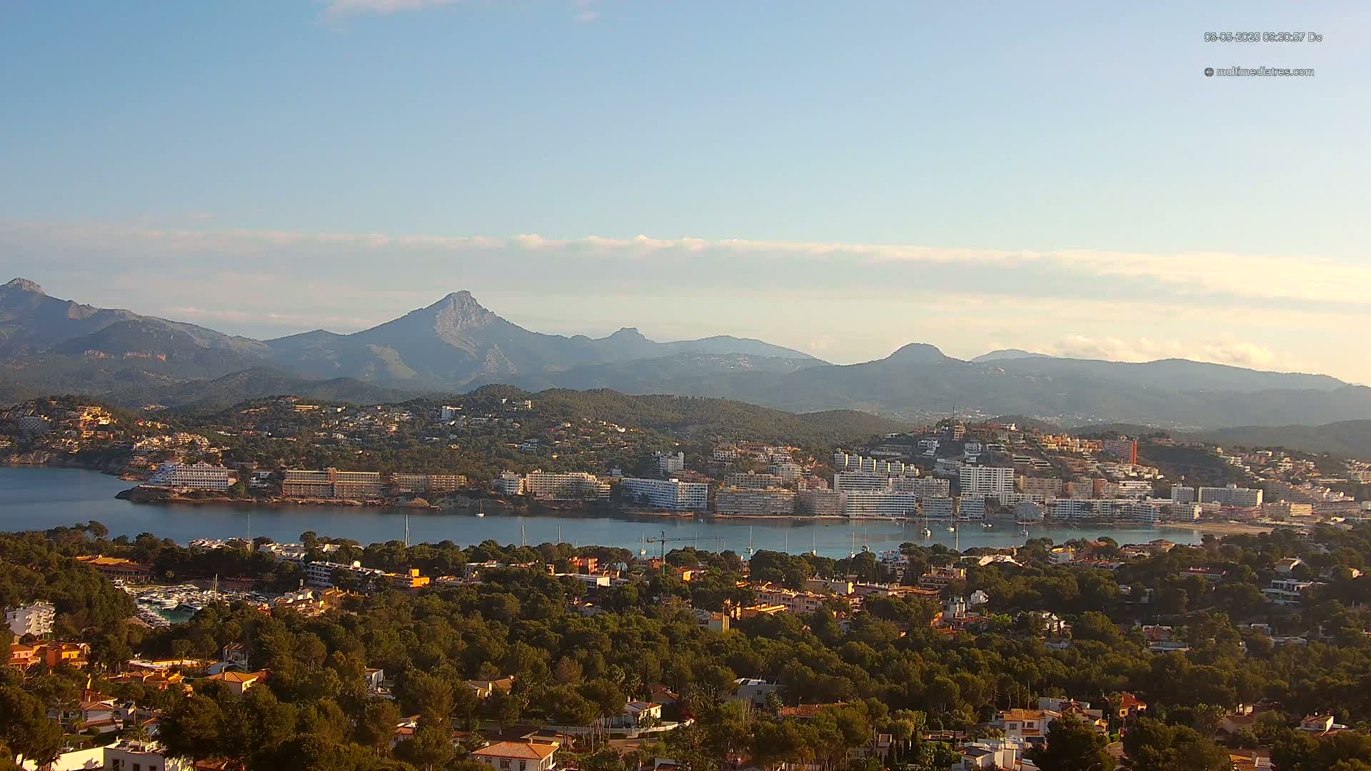 A coastal town nestled amongst green hills and mountains under a mostly clear sky.