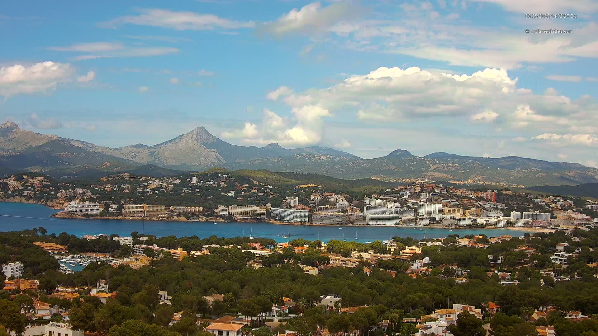 A coastal town nestled between a bay and a mountain range under a partly cloudy sky.