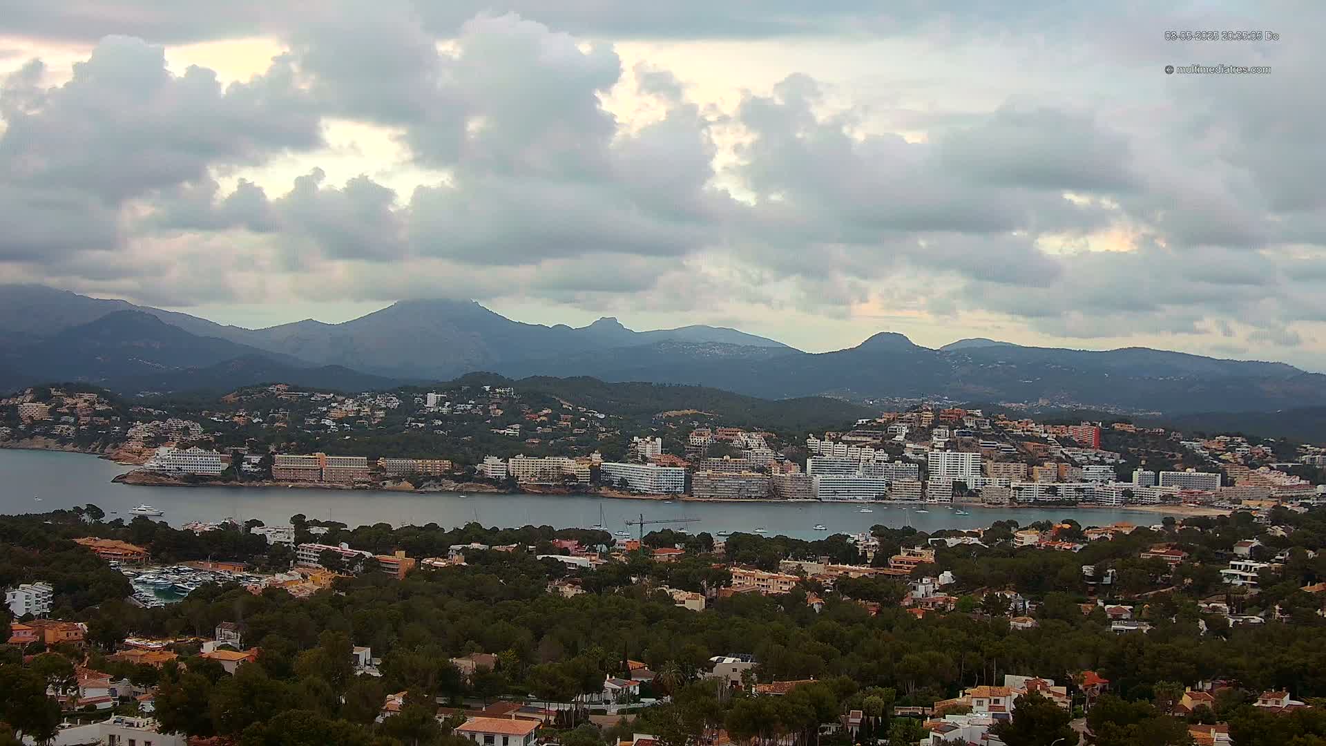 A coastal town nestled between a bay and a mountain range under a cloudy sky.