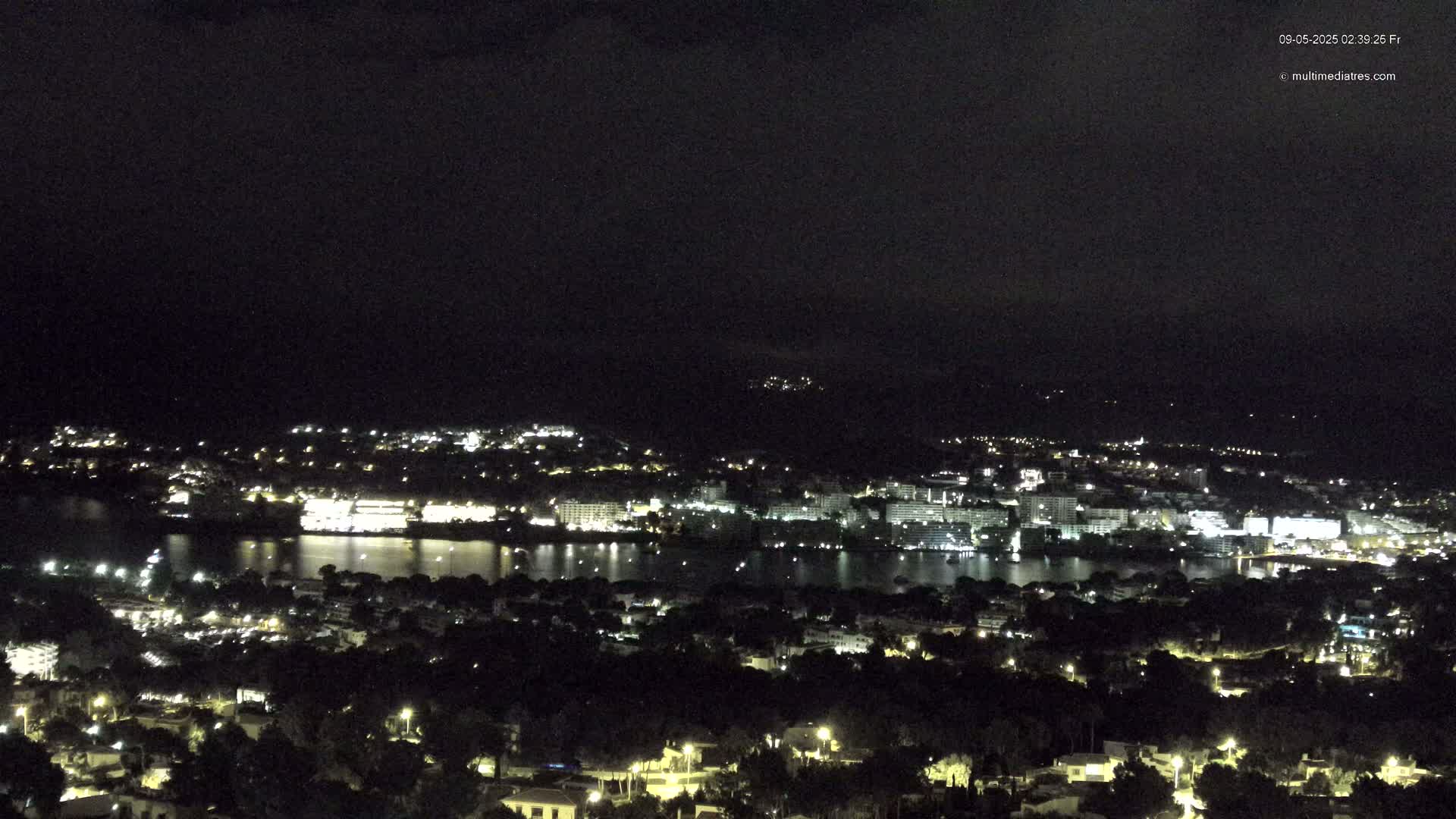 A nighttime, aerial view shows a city with many lights spread across a body of water, surrounded by darker land and hills.