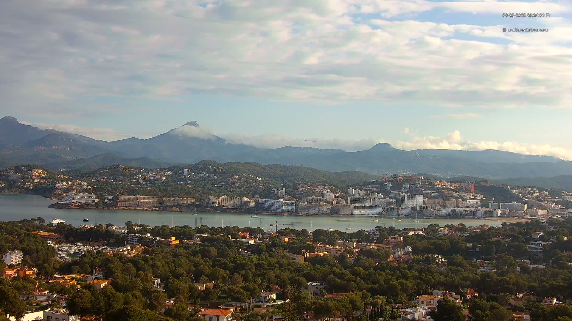 A coastal town nestled amongst green hills and mountains under a partly cloudy sky.