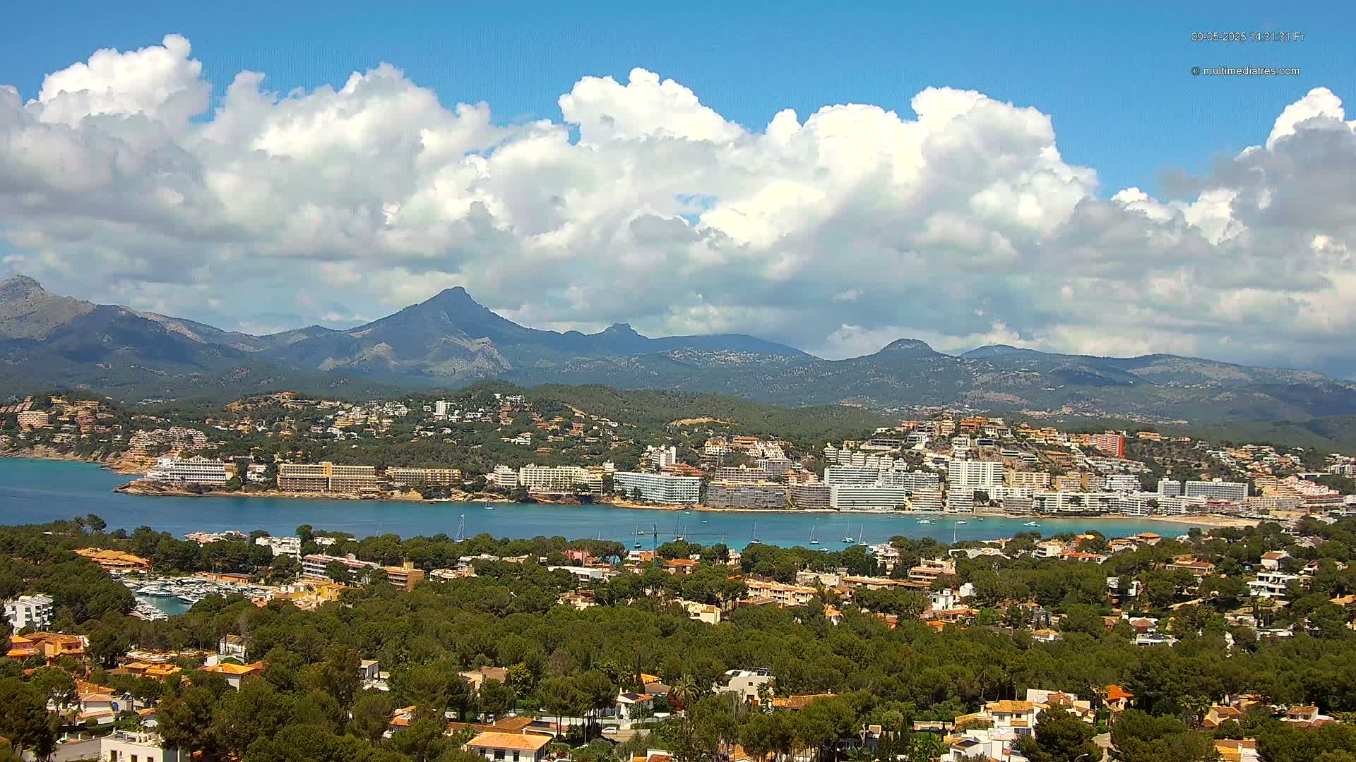 A coastal town nestled amongst green hills and a calm bay, backed by a range of mountains under a partly cloudy sky.