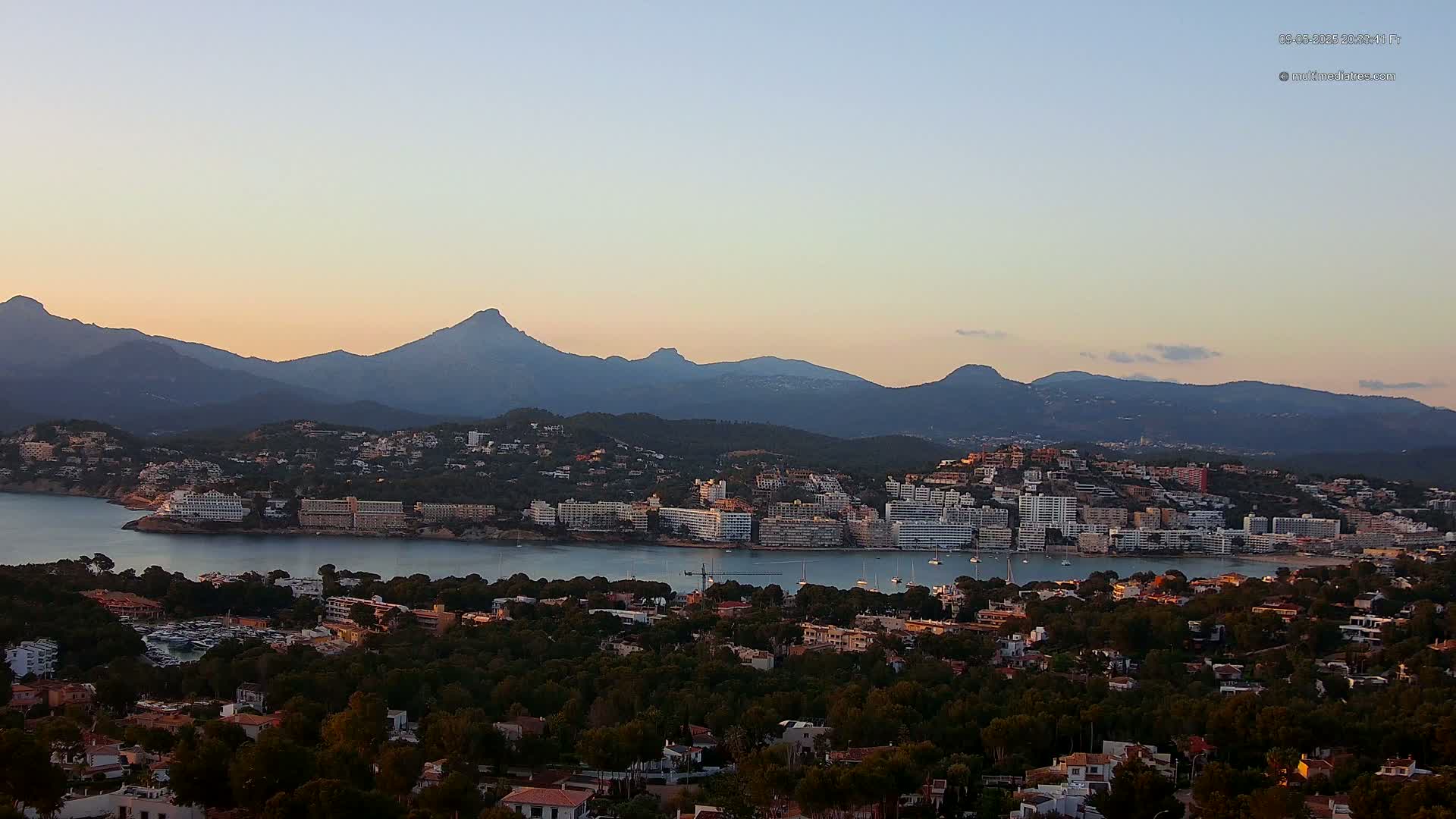 A coastal town nestled in a bay, backed by mountains, is bathed in the soft light of either sunrise or sunset.