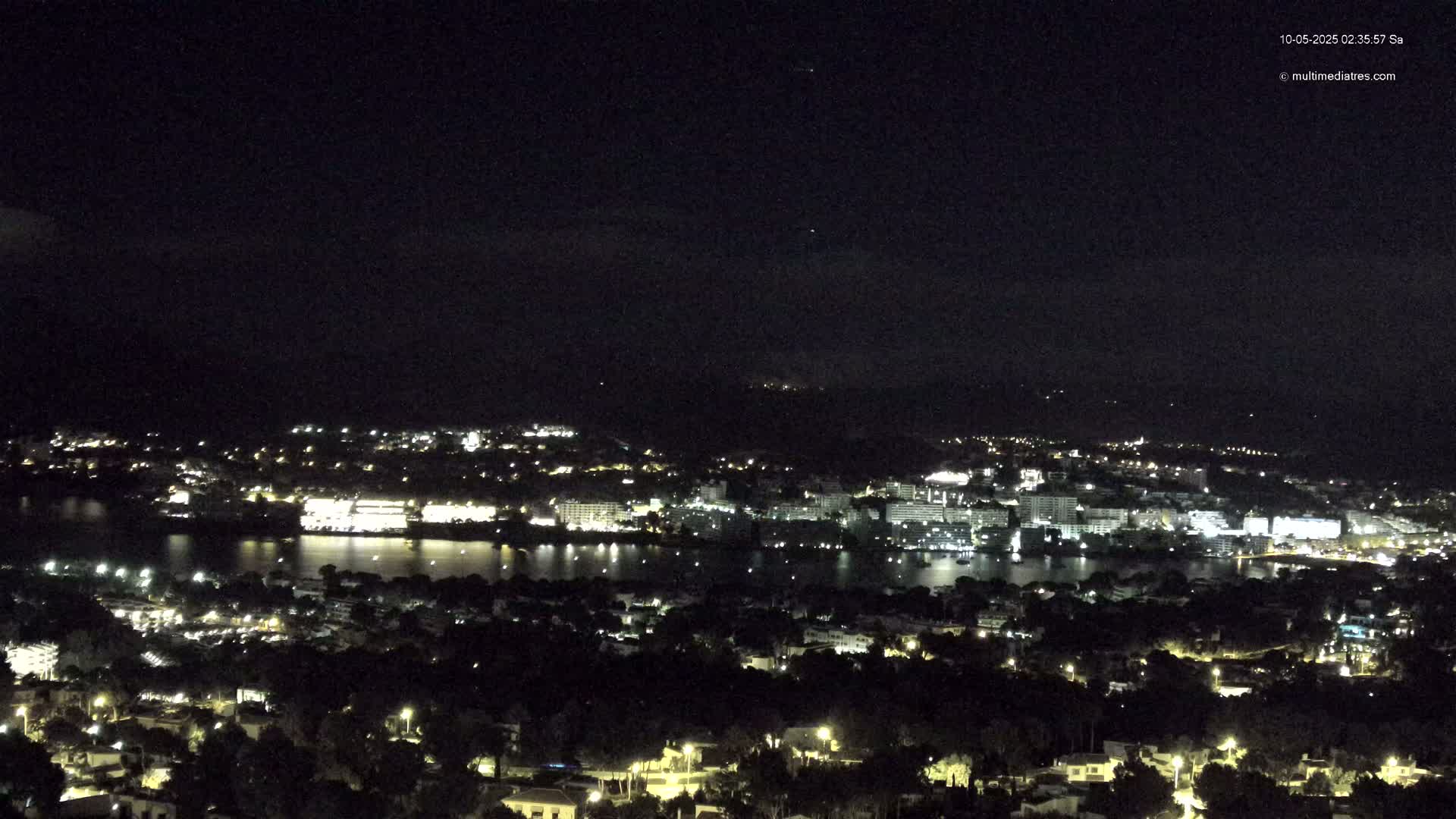 A nighttime view of a coastal city with its lights illuminating the buildings and surrounding water under a clear, dark sky.