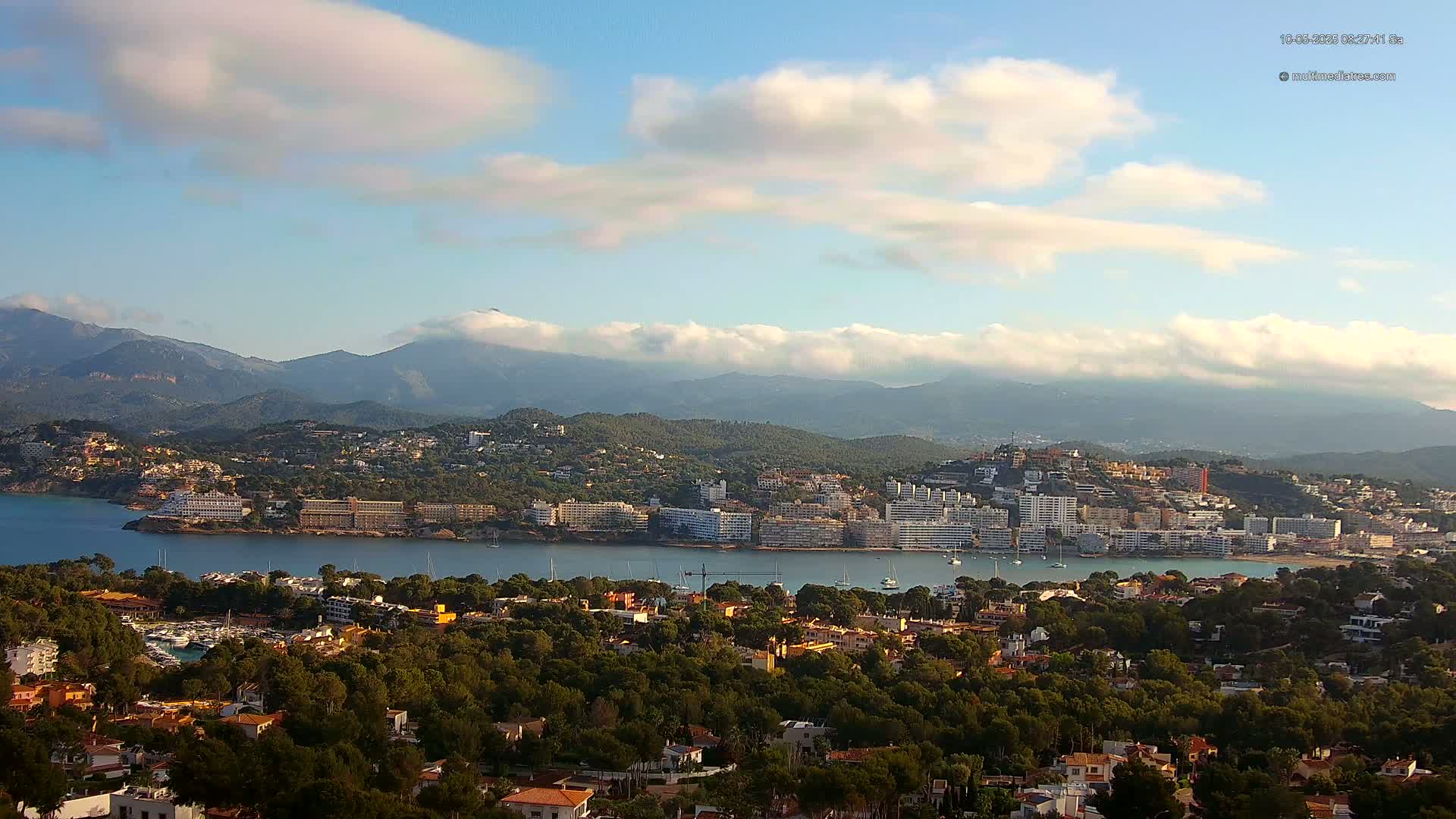 A coastal town nestled between a calm bay and a mountain range under a partly cloudy sky.