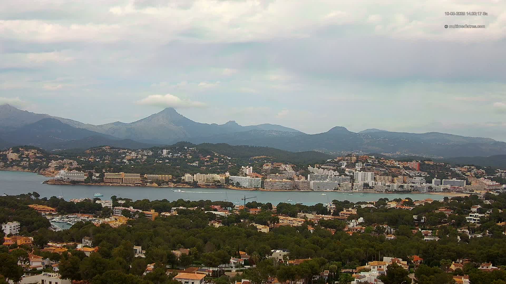 A coastal town nestled among green hills and mountains under a cloudy sky, with a body of water separating the town from a larger collection of buildings along the coast.
