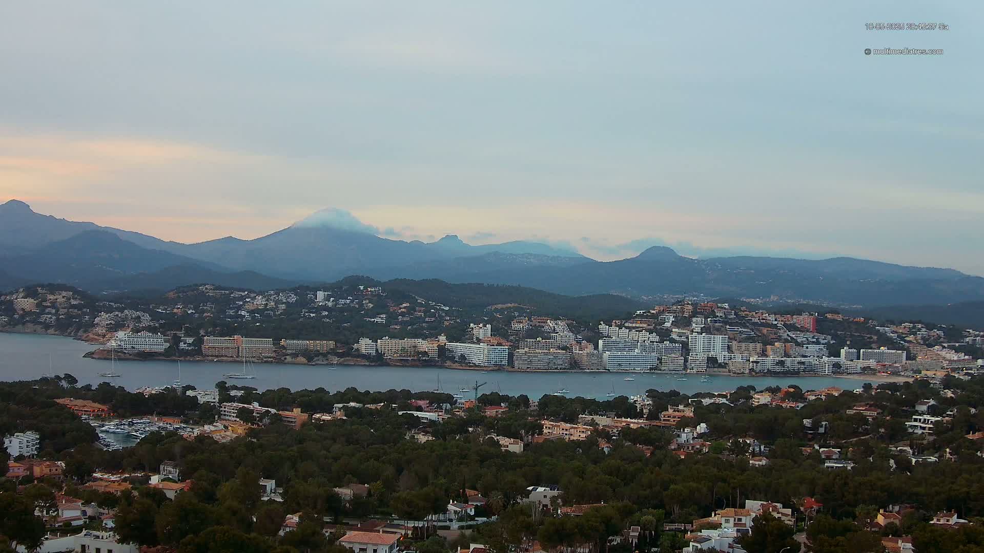 A coastal town nestled between a calm bay and a mountain range under a mostly cloudy sky.