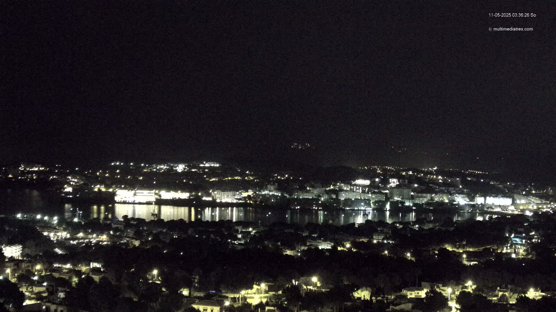 A nighttime view of a city and body of water, with the city lights reflecting on the water under a clear, dark sky.