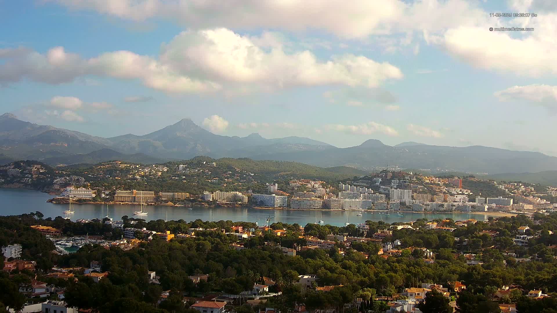A coastal town nestled among green hills and mountains under a partly cloudy sky, with boats in the bay.