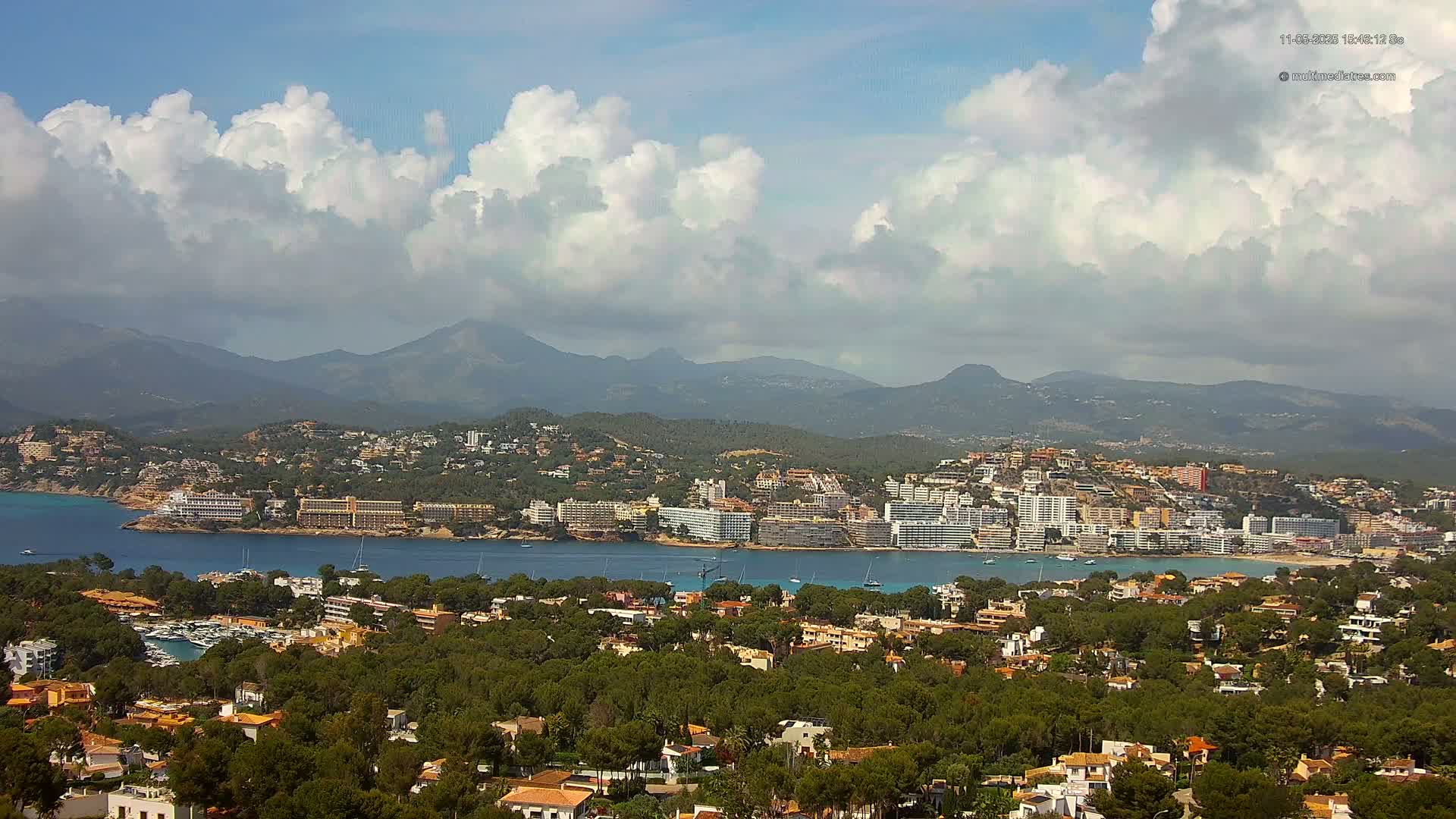 A coastal town nestled amongst green hills and mountains under a partly cloudy sky, with a calm body of water separating the town from a larger bay area.