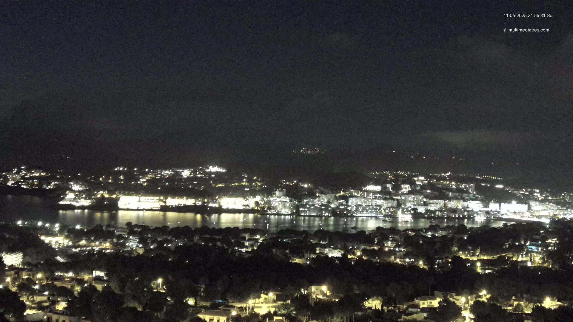 A nighttime, high-angle view shows a city illuminated along a dark body of water, nestled between hills under a clear, dark sky.