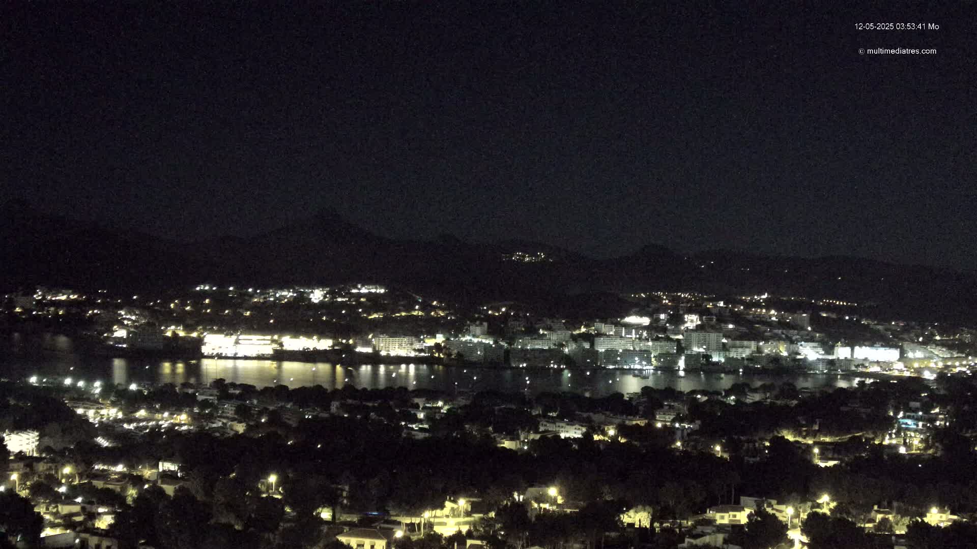 A nighttime view shows a city and body of water illuminated by lights, nestled at the base of dark mountains under a clear sky.