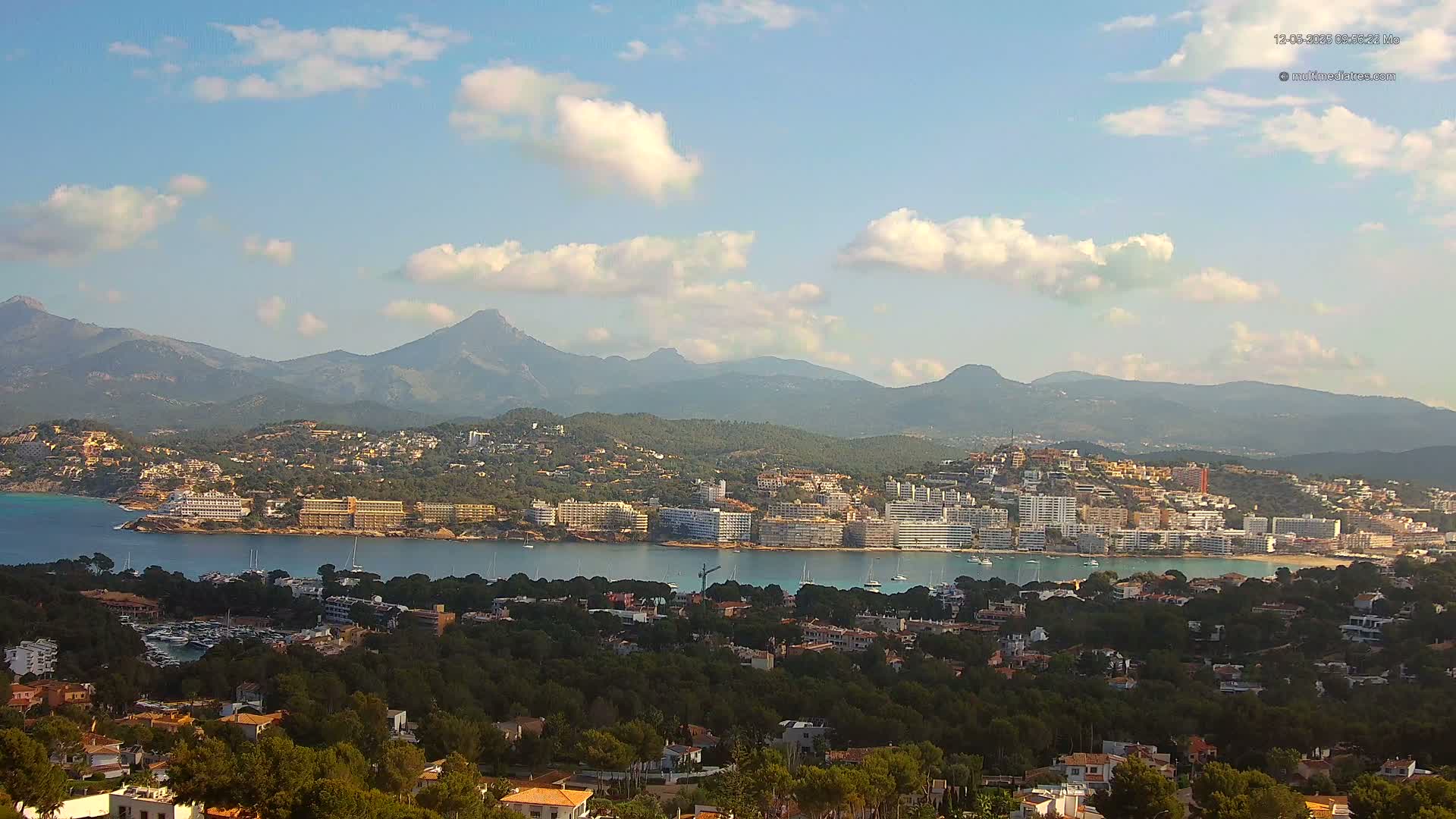 A coastal town nestled between a calm bay and a mountain range under a partly cloudy sky.