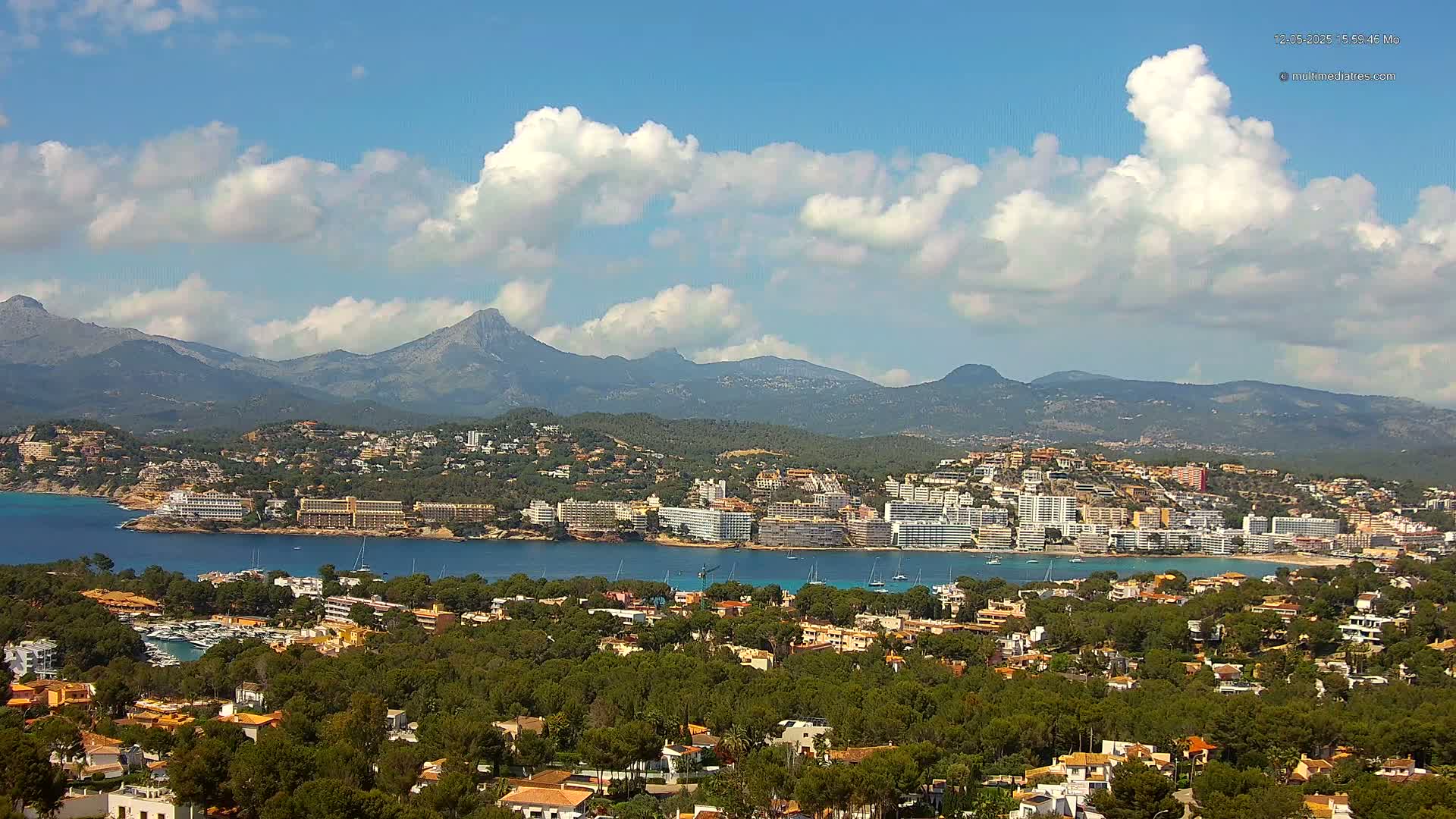A coastal town nestled amongst green hills and mountains under a partly cloudy sky, with a calm body of water separating the town from the mountains.