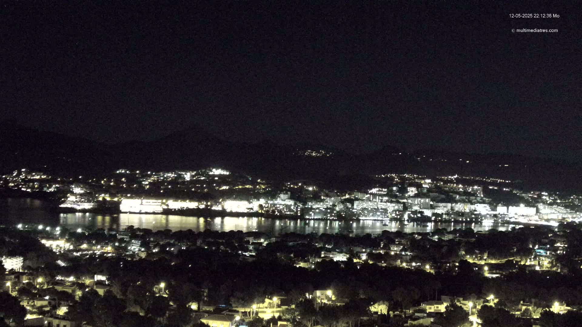 A nighttime view of a city illuminated by lights across a body of water, with dark mountains in the background under a clear sky.