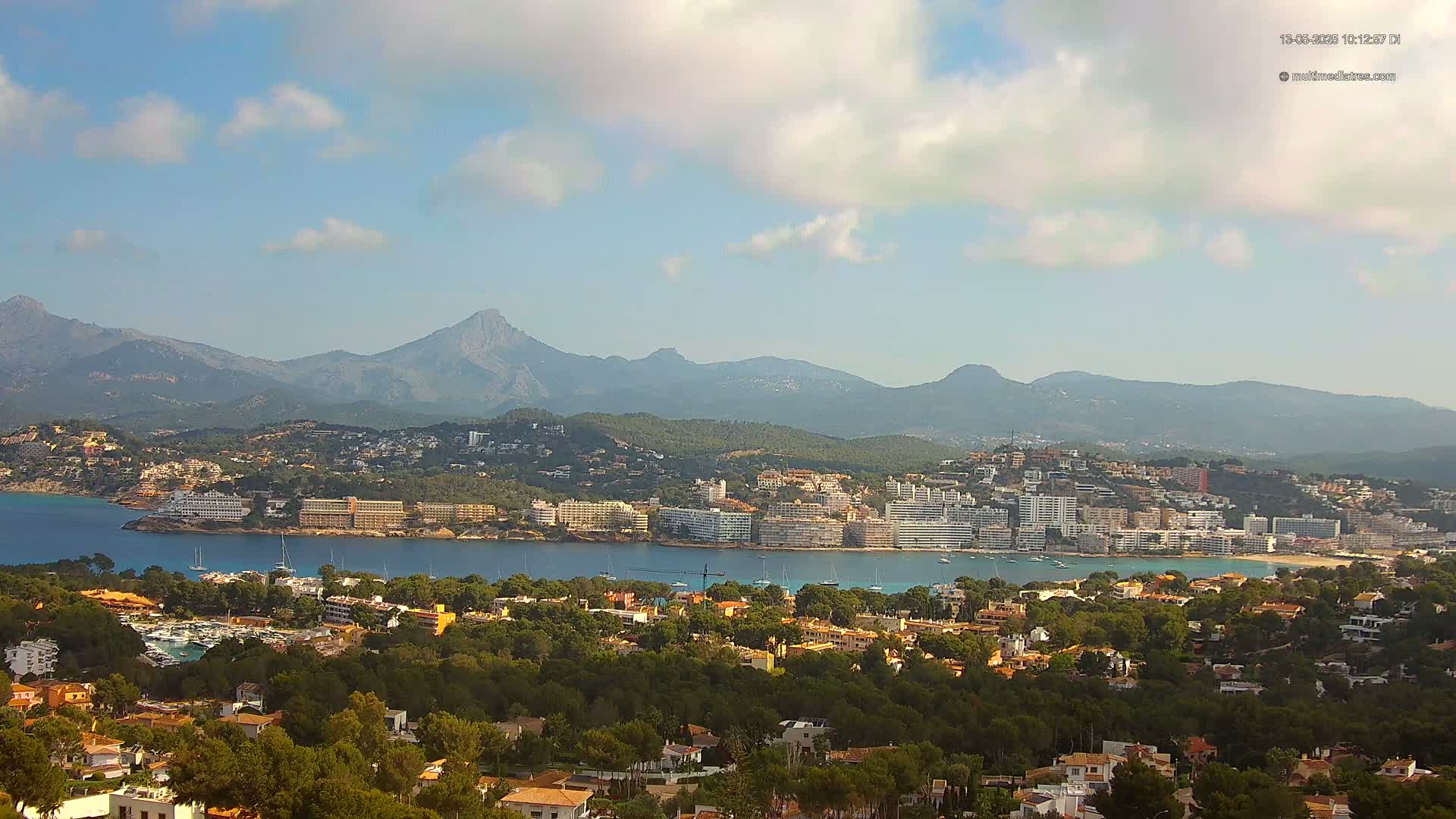 A coastal town nestled in a bay, backed by mountains under a partly cloudy sky.