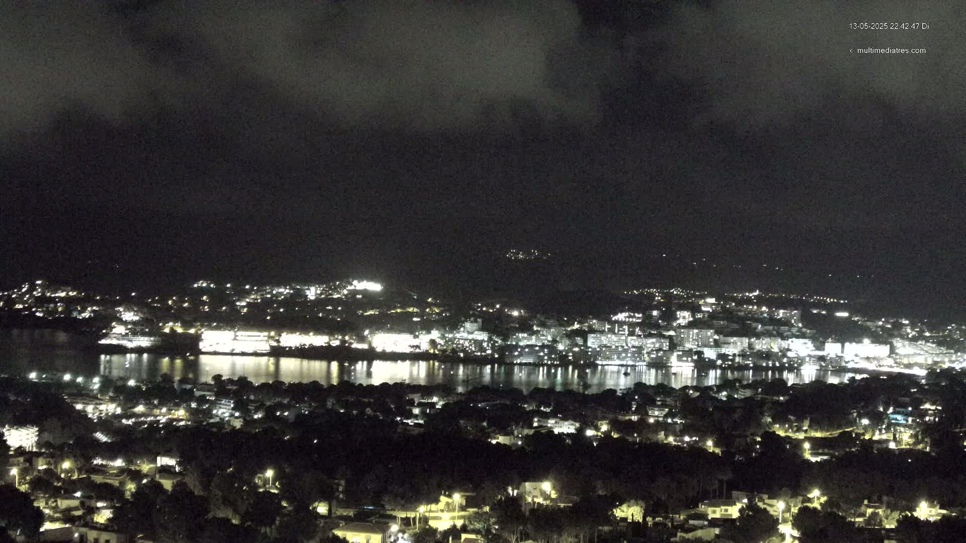 A nighttime, overcast view of a city and its harbor illuminated by lights, reflecting on the calm water.