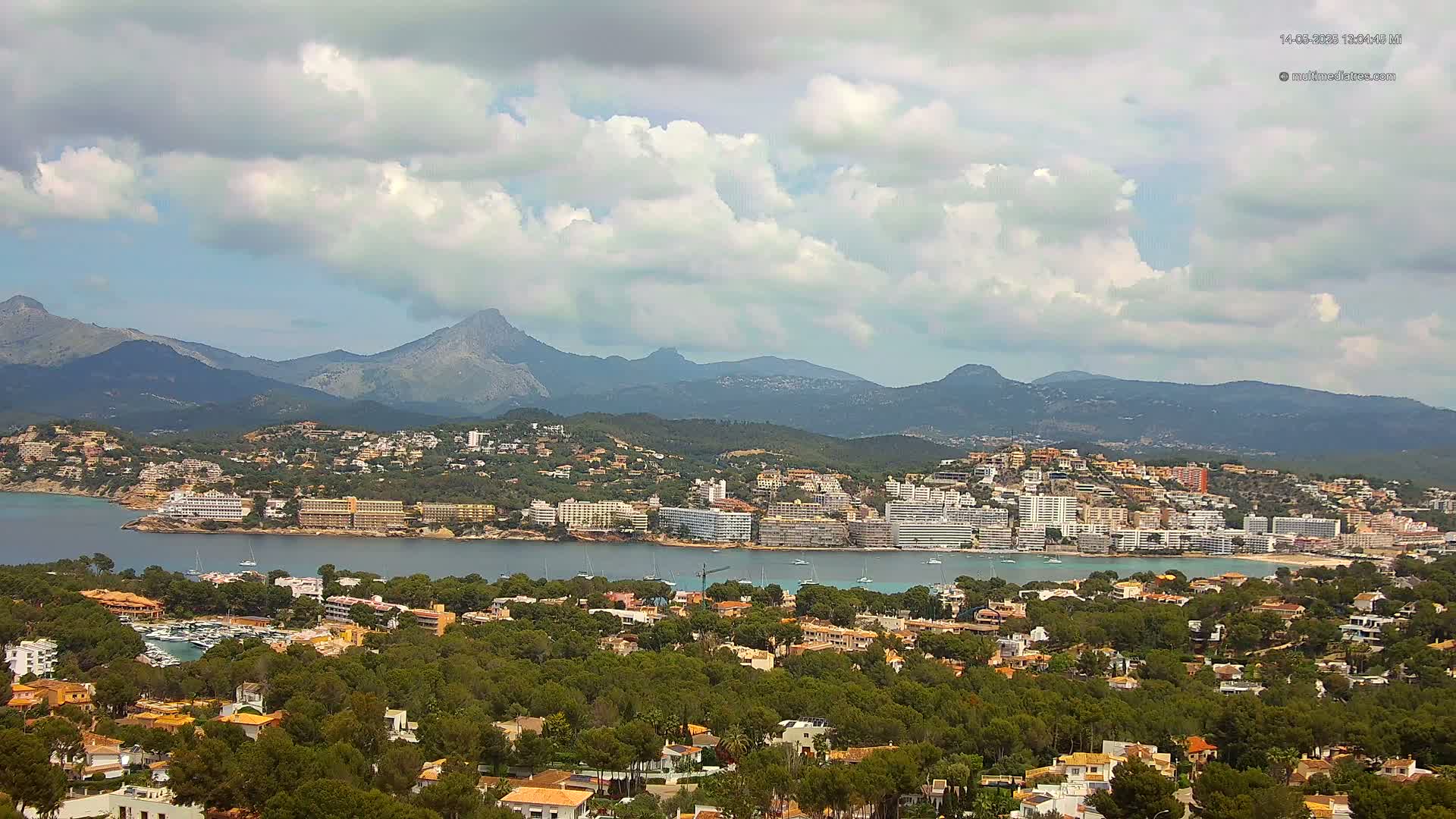 A coastal town nestled among green hills and mountains under a partly cloudy sky, with a calm body of water separating the town from a larger mountainous region.