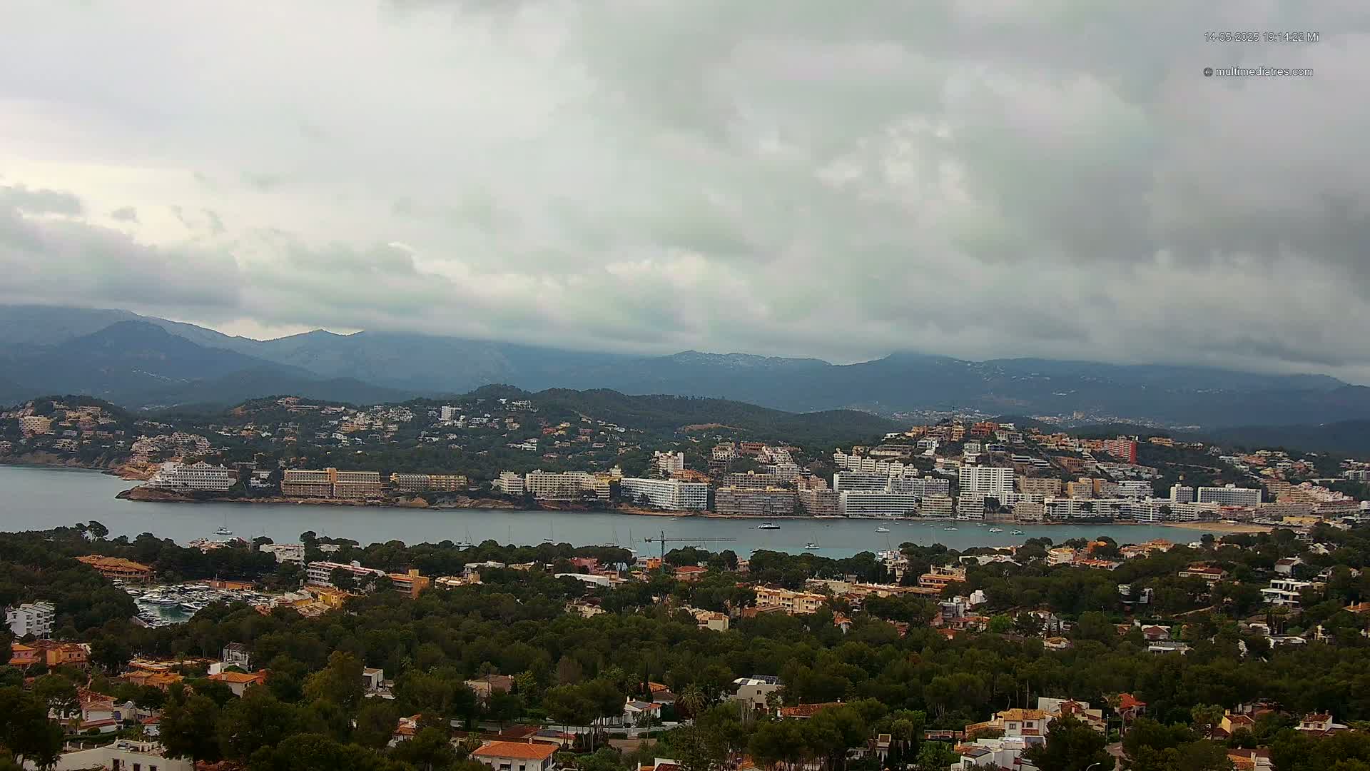 A coastal town nestled between a calm bay and a mountain range under an overcast sky.