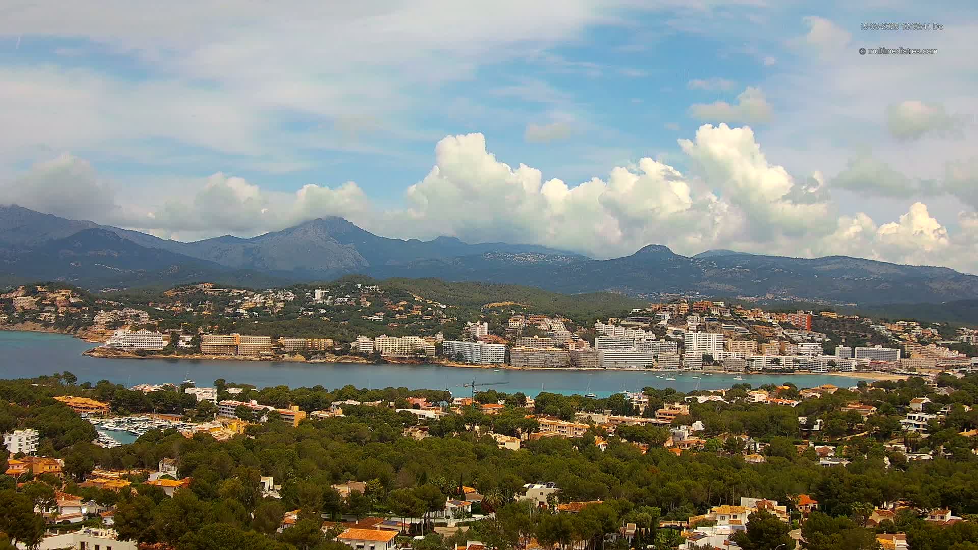 A coastal town nestled between a bay and a mountain range under a partly cloudy sky.