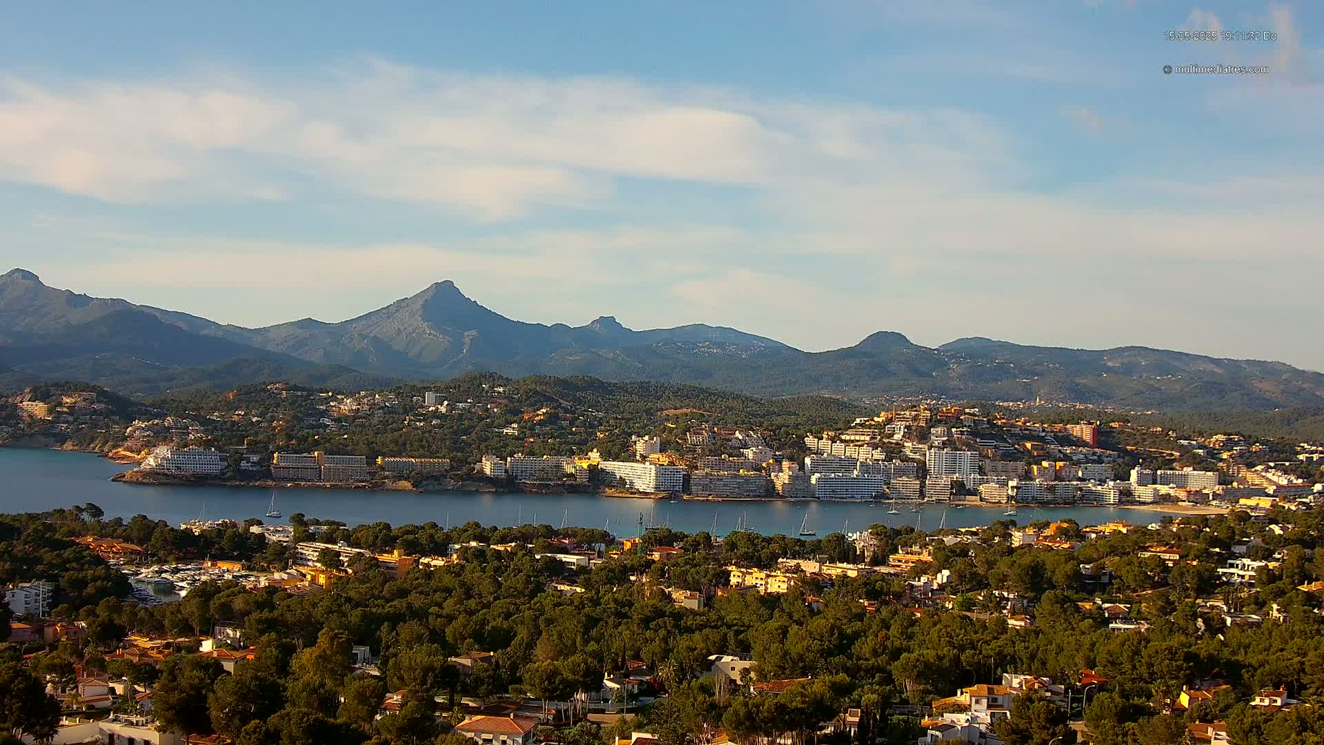 A coastal town nestled among green hills and mountains, with a calm bay and clear skies.