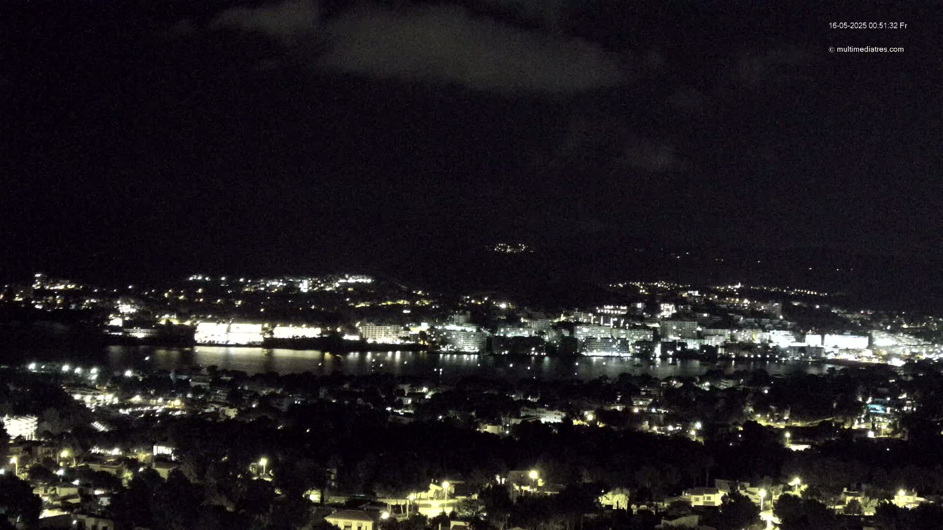 A nighttime view of a city illuminated by lights, situated beside a body of water under a partly cloudy sky.