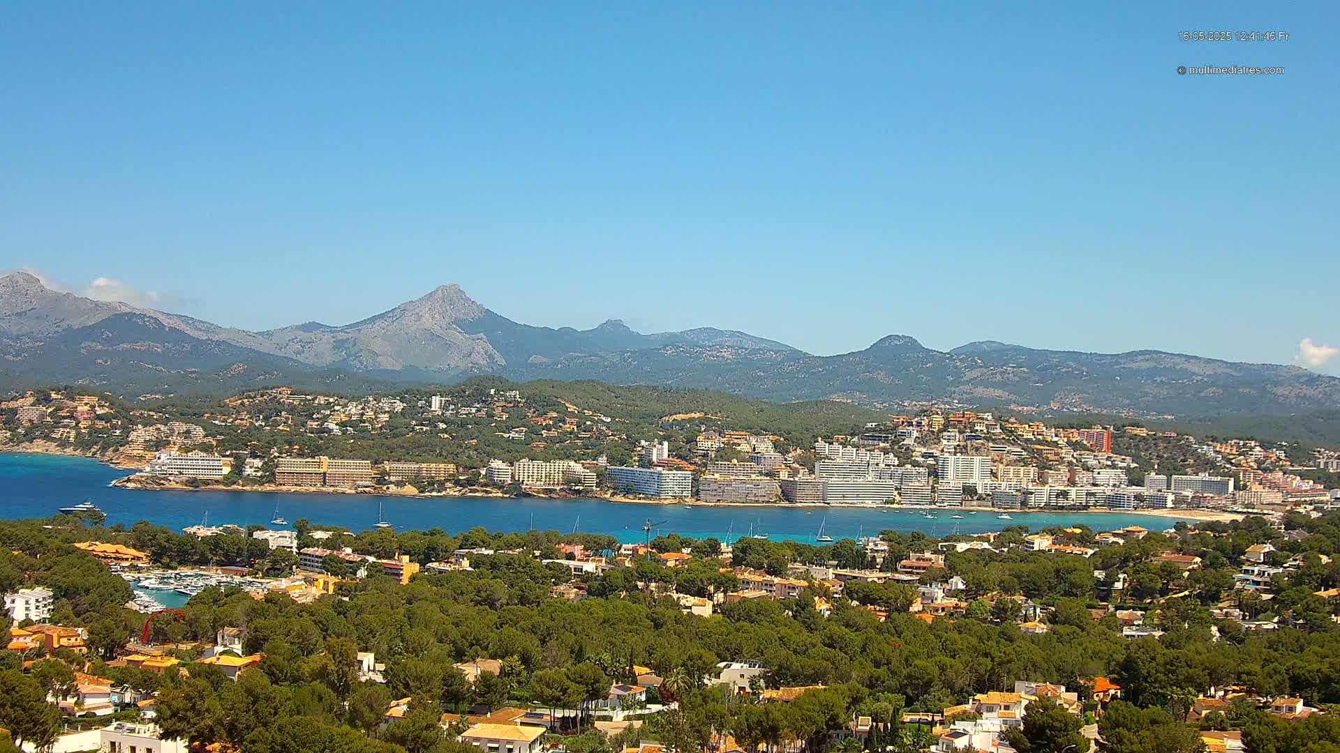 A coastal town nestled amongst green hills and mountains under a clear blue sky overlooks a calm blue bay dotted with boats.