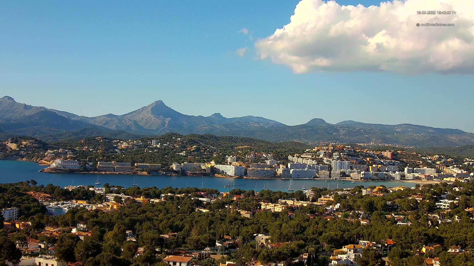 A coastal town nestled between a calm bay and a mountain range under a mostly sunny sky with a single puffy cloud.