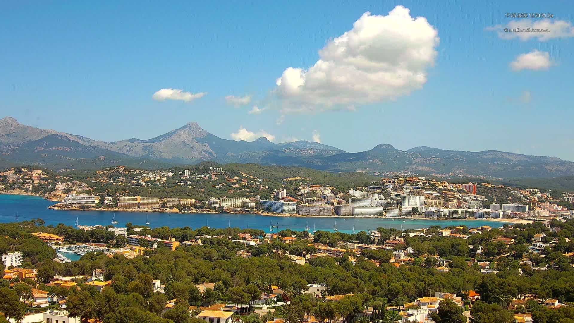 A coastal town nestled in a bay, backed by lush green hills and a mountain range under a mostly sunny sky with a few puffy clouds.