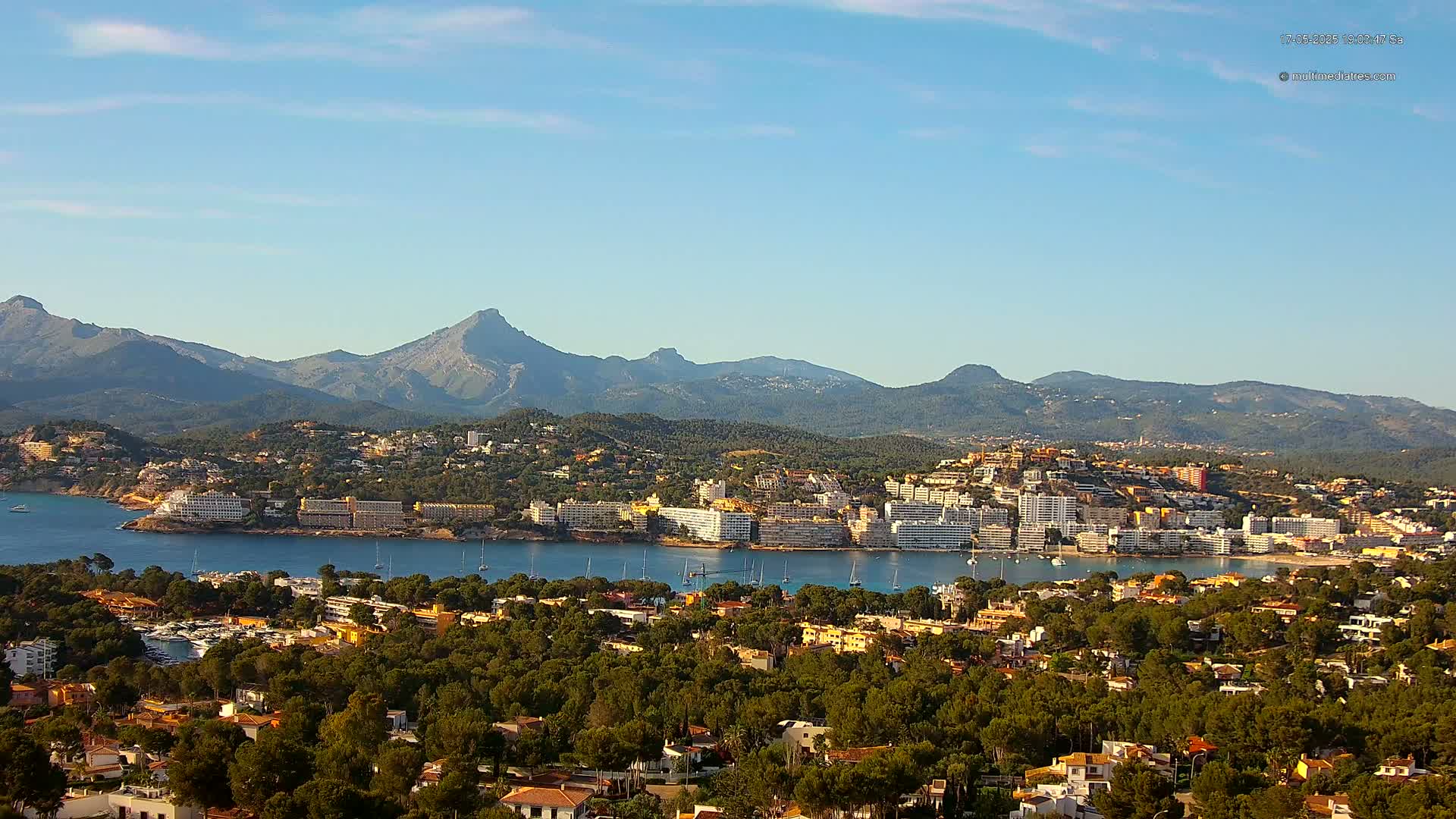 A coastal town nestled between a calm bay and a mountain range under a mostly clear sky.