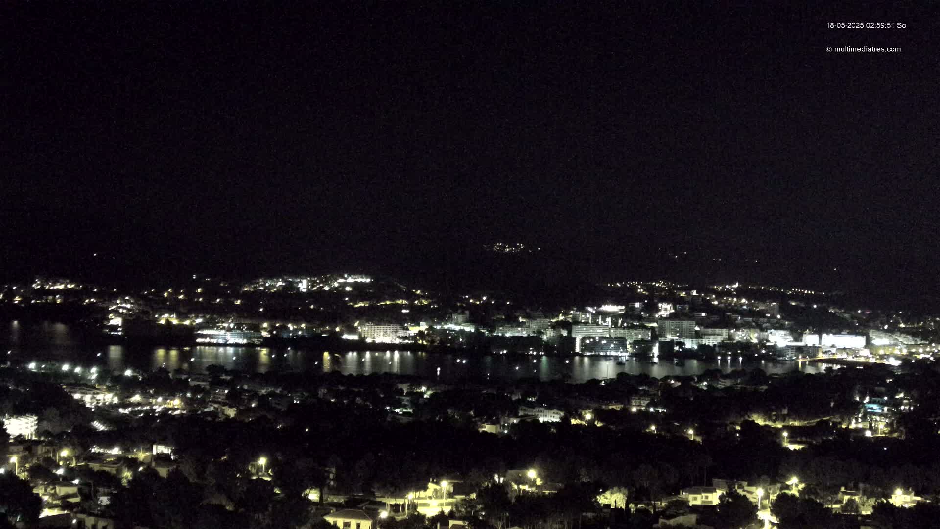 A nighttime view of a coastal city illuminated by lights, reflecting on a calm body of water under a clear, dark sky.