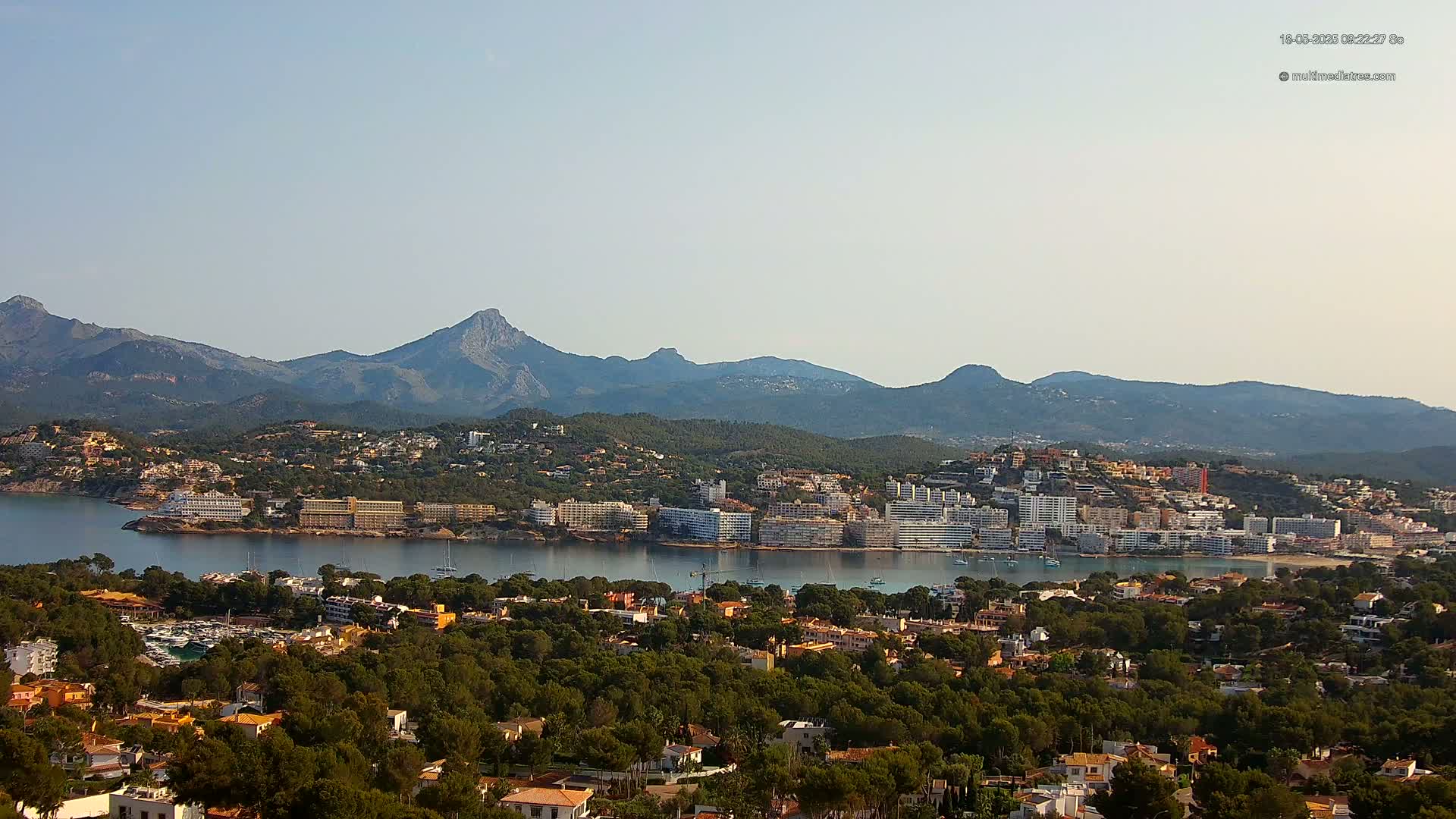 A coastal town nestled amongst green hills and mountains under a clear sky, with a calm body of water separating the town from a peninsula with large buildings.