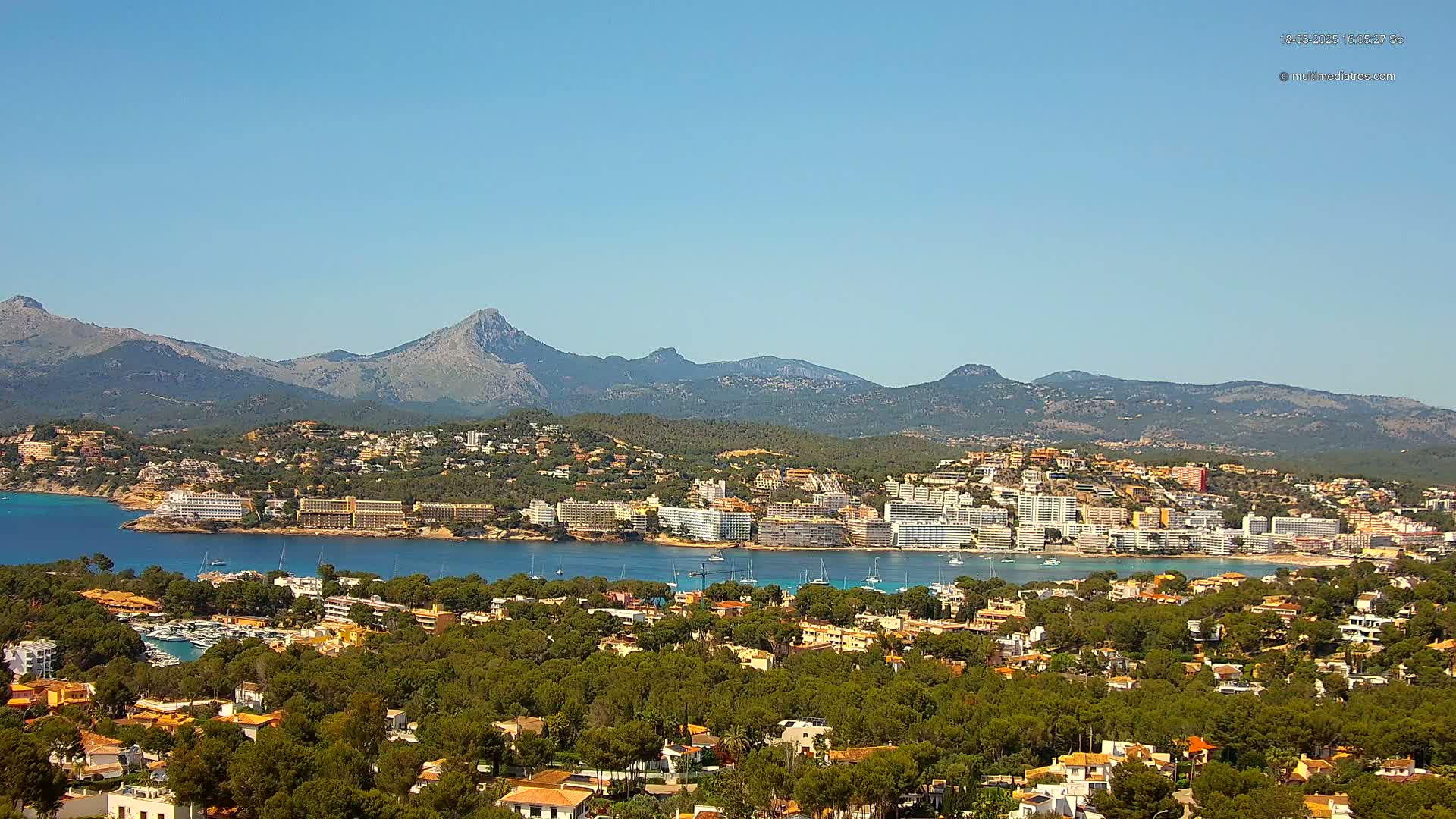 A coastal town nestled amongst green hills and mountains, overlooking a calm bay on a clear, sunny day.