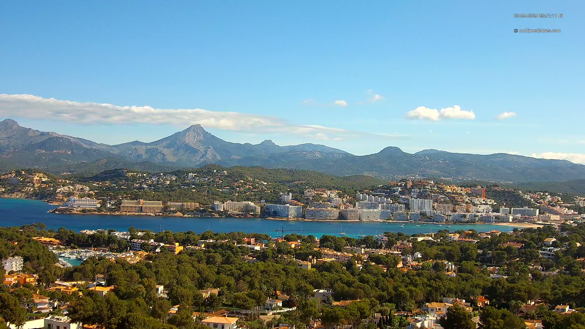 A coastal town nestled among green hills and mountains under a mostly clear blue sky with a few scattered clouds.
