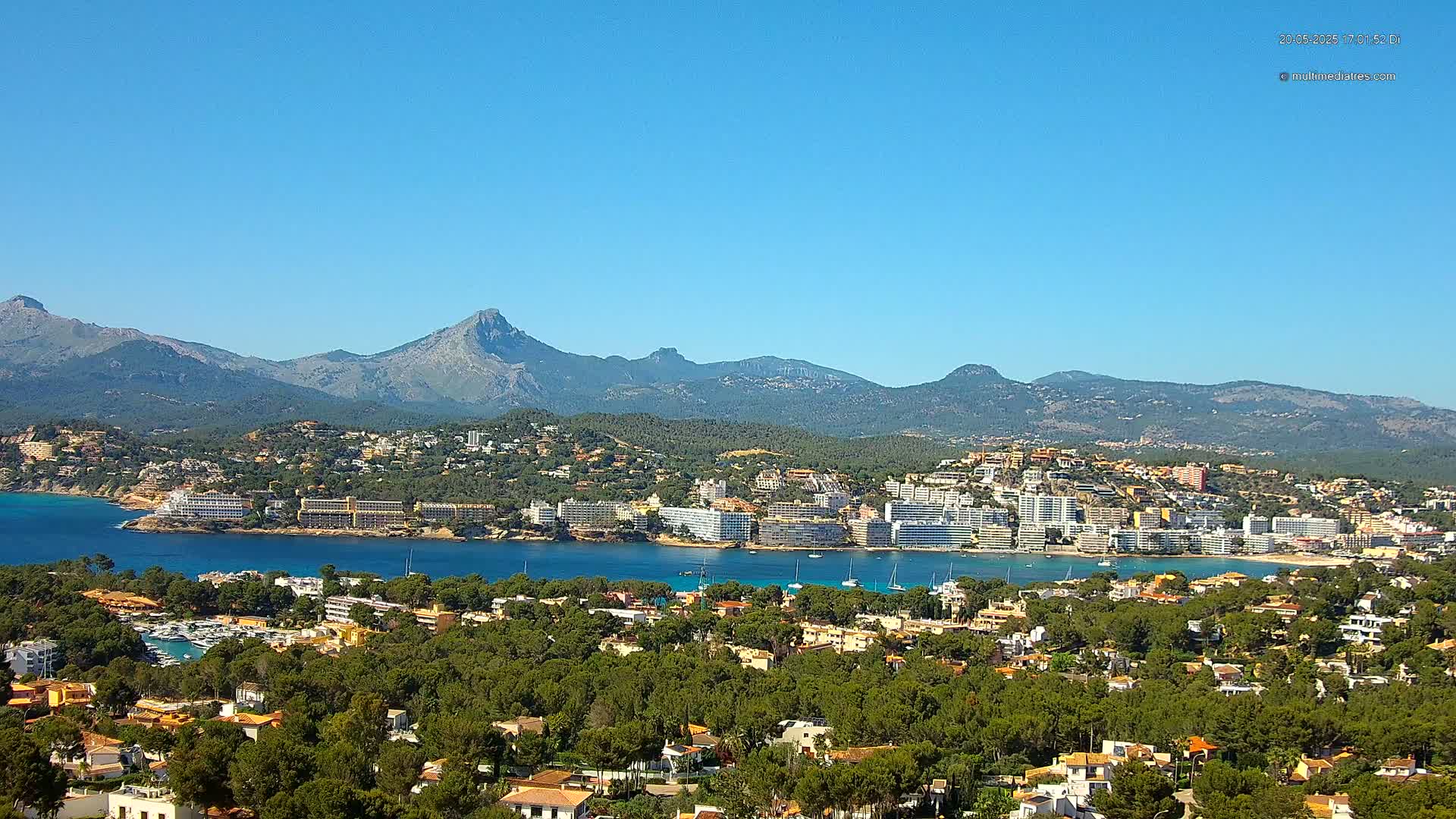 A coastal town nestled amongst green hills and mountains under a clear blue sky.