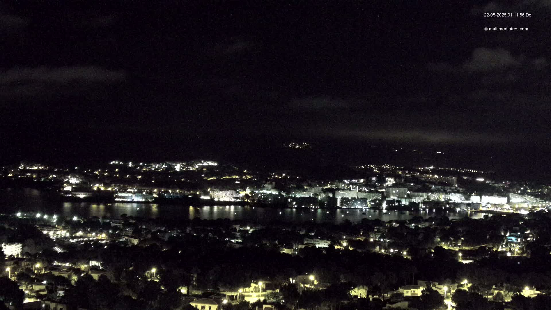 A nighttime view of a city and body of water, under a partly cloudy sky, shows numerous lights illuminating the cityscape and its reflection on the water.