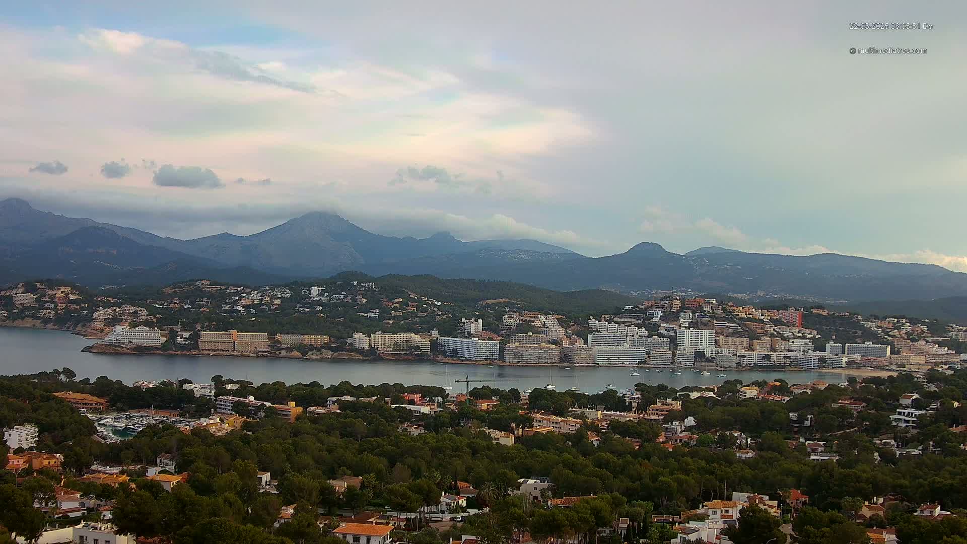 A coastal town nestled between a calm bay and a mountain range under a partly cloudy sky.