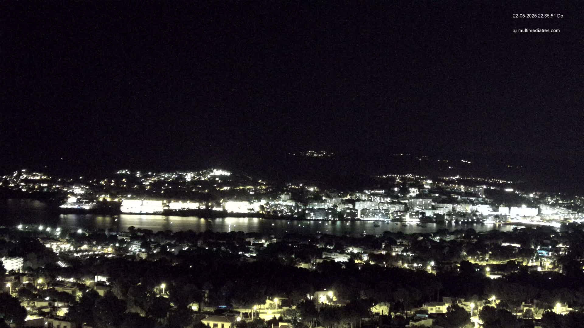 A nighttime aerial view shows a city illuminated by lights, situated beside a body of water, under a clear, dark sky.