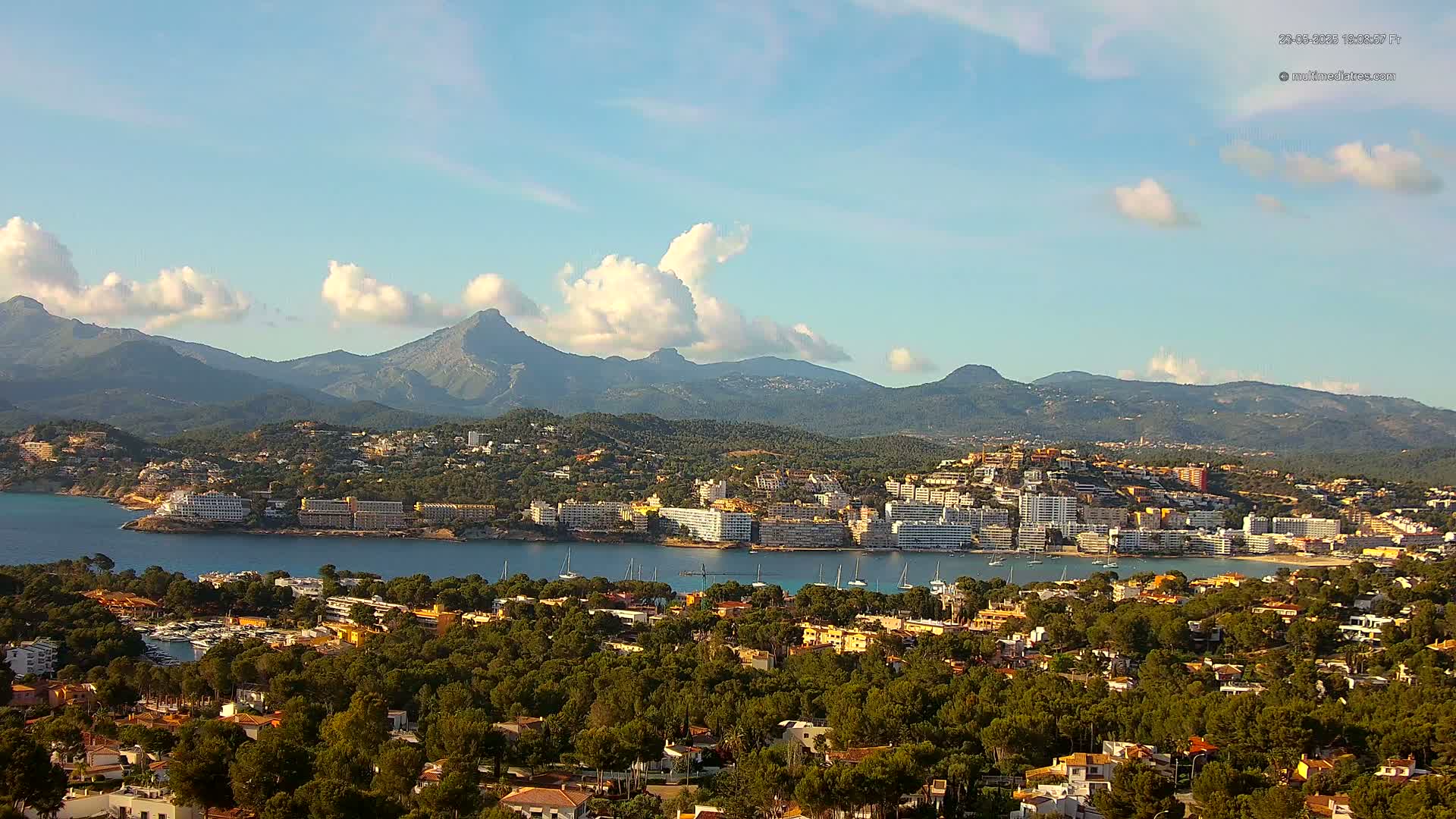 A coastal town nestled amongst lush green trees and hills, with a calm bay of water and a mountainous backdrop under a partly cloudy sky.