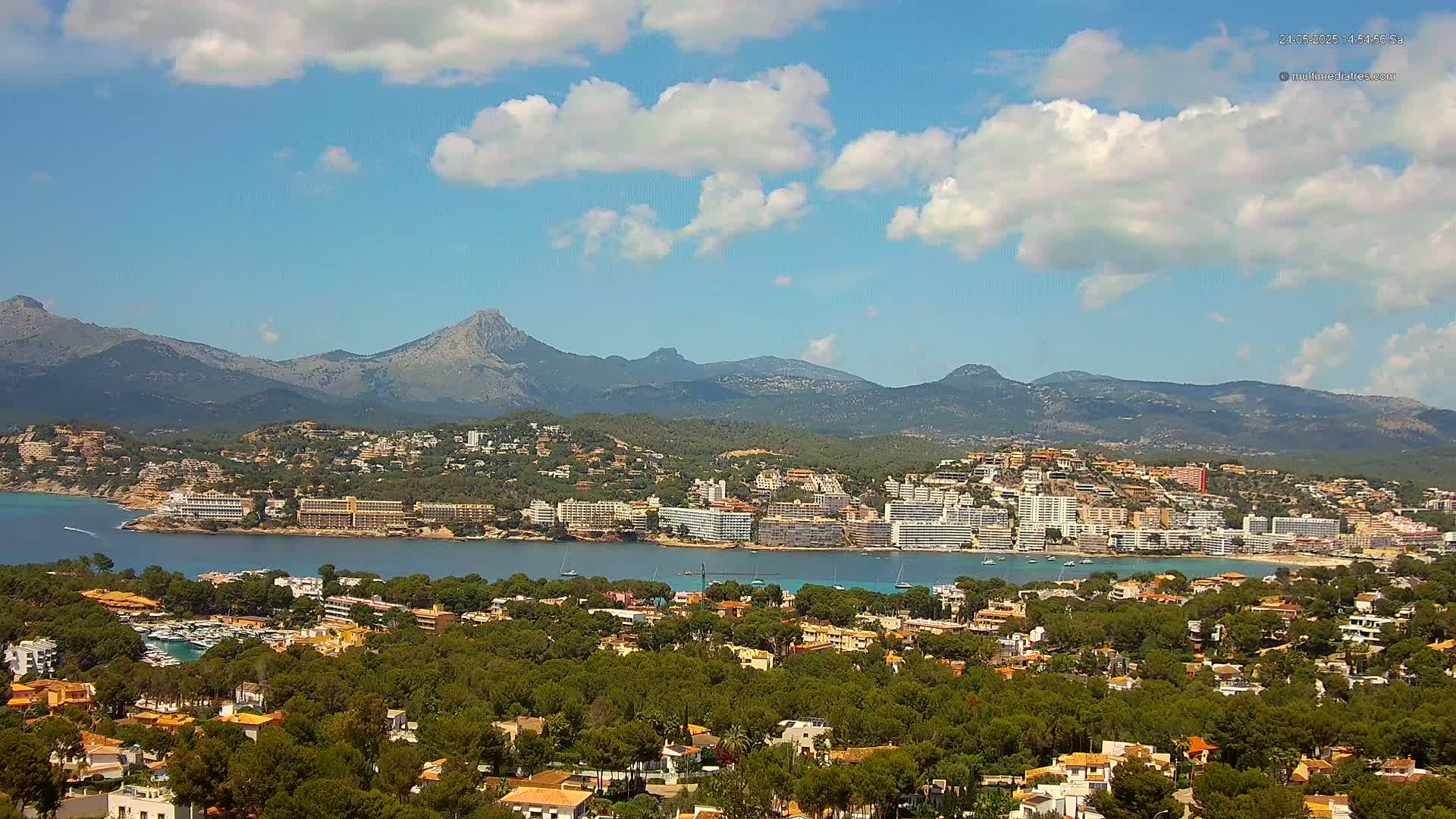 A coastal town with numerous buildings and hotels nestled among green trees, backed by a range of mountains under a partly cloudy sky.
