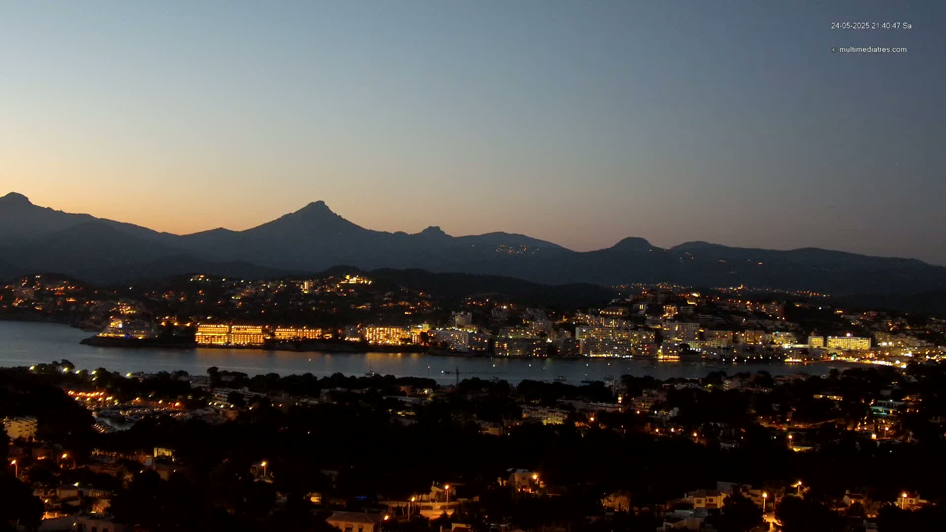 A coastal town illuminated at night, nestled between a calm body of water and a dark mountain range under a twilight sky.