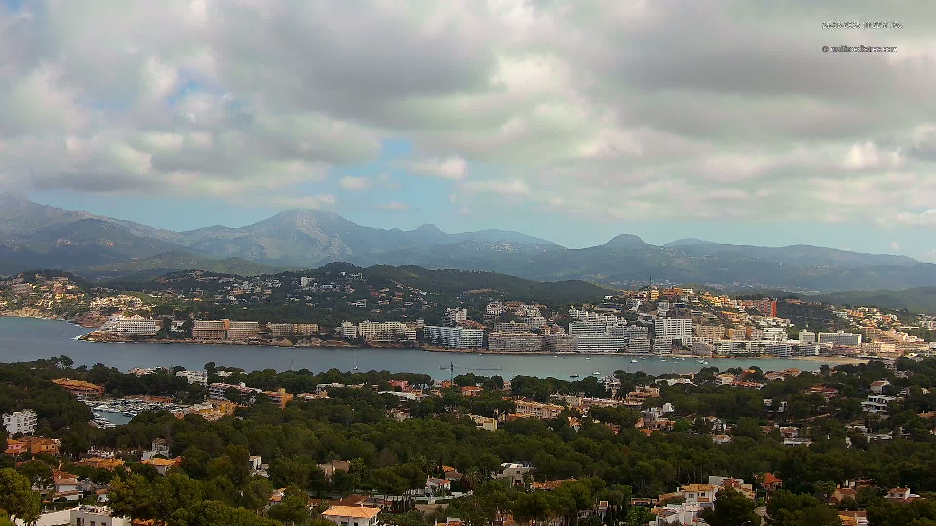 A coastal town nestled between a calm bay and a mountain range under a partly cloudy sky.