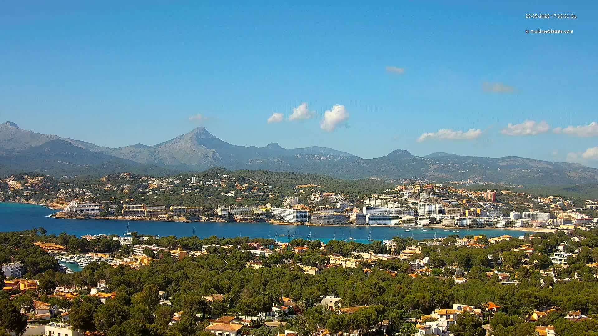 A coastal town nestled amongst green hills and a calm blue bay, backed by a mountain range under a mostly clear blue sky with a few scattered clouds.