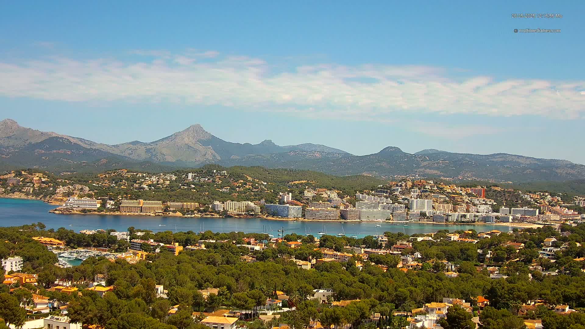 A coastal town nestled amongst lush green hills and mountains under a mostly sunny sky with scattered clouds.