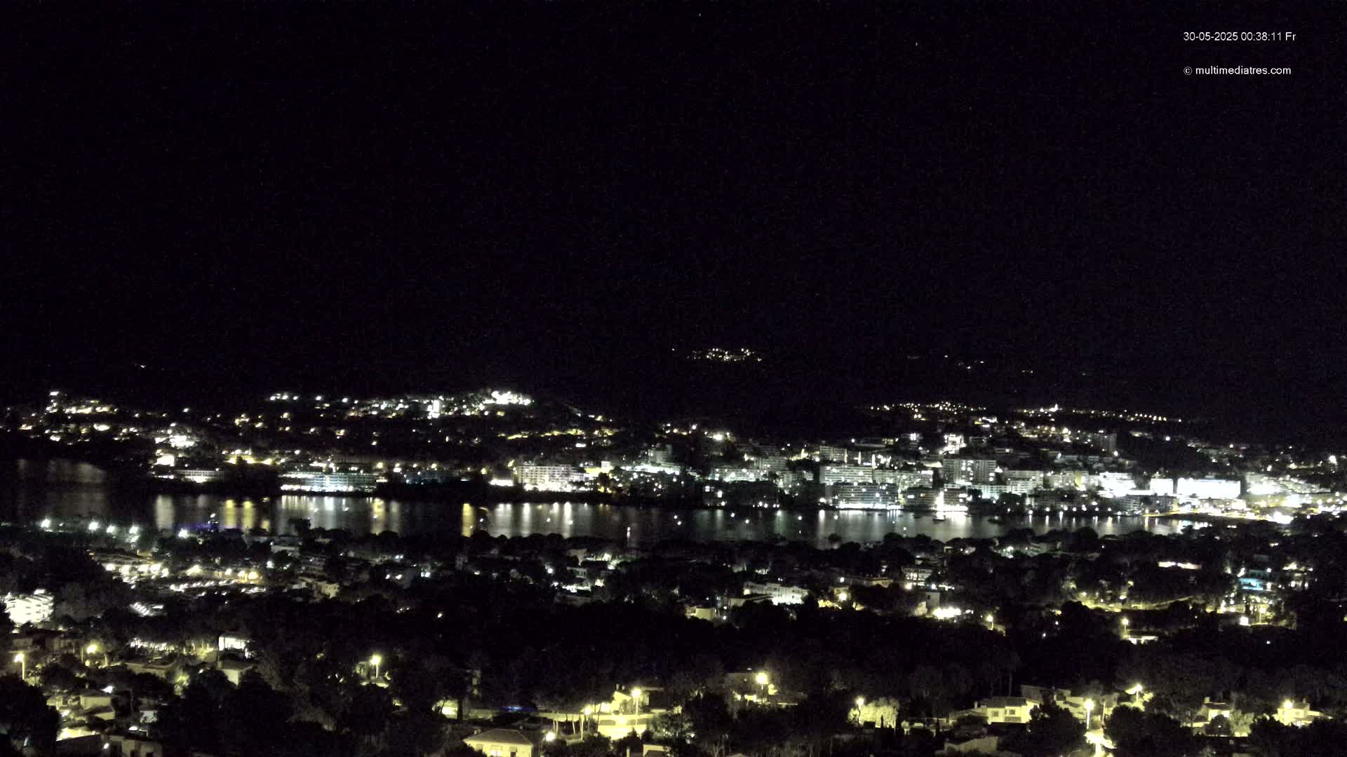 A nighttime view of a coastal town illuminated with lights, situated next to a body of water reflecting the city lights under a clear, dark sky.