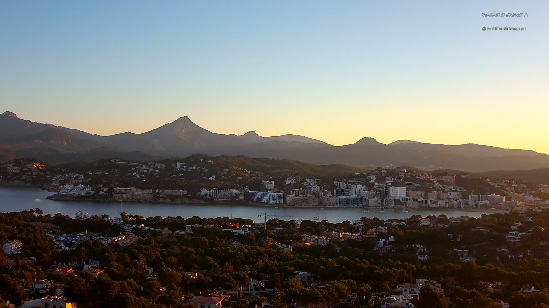 A coastal town nestled beside a calm bay, backed by a range of mountains under a clear, sunset sky.