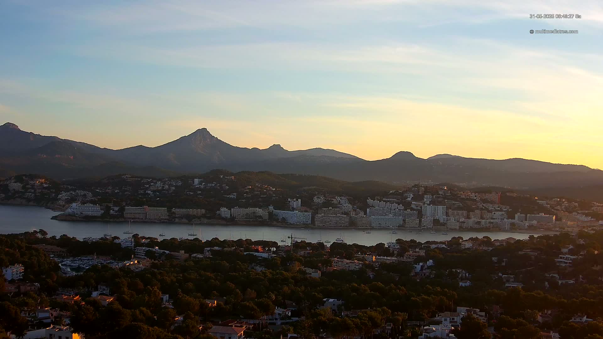 A coastal town nestled among a bay, with numerous buildings and boats, sits at the foot of a mountain range under a clear, sunset sky.